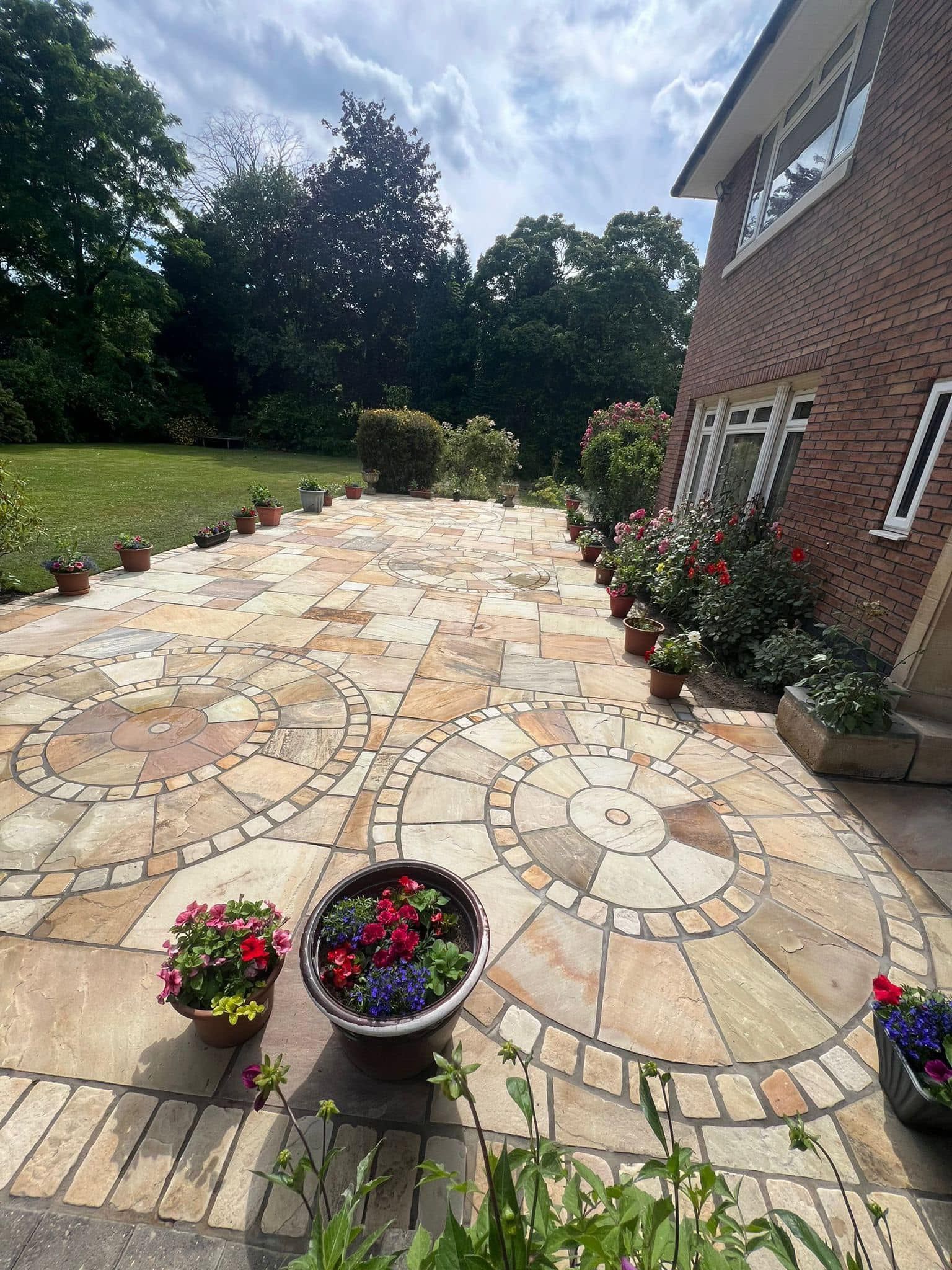 Stone patio with flower pots lining the edges, and a brick house in the background under a cloudy sky.