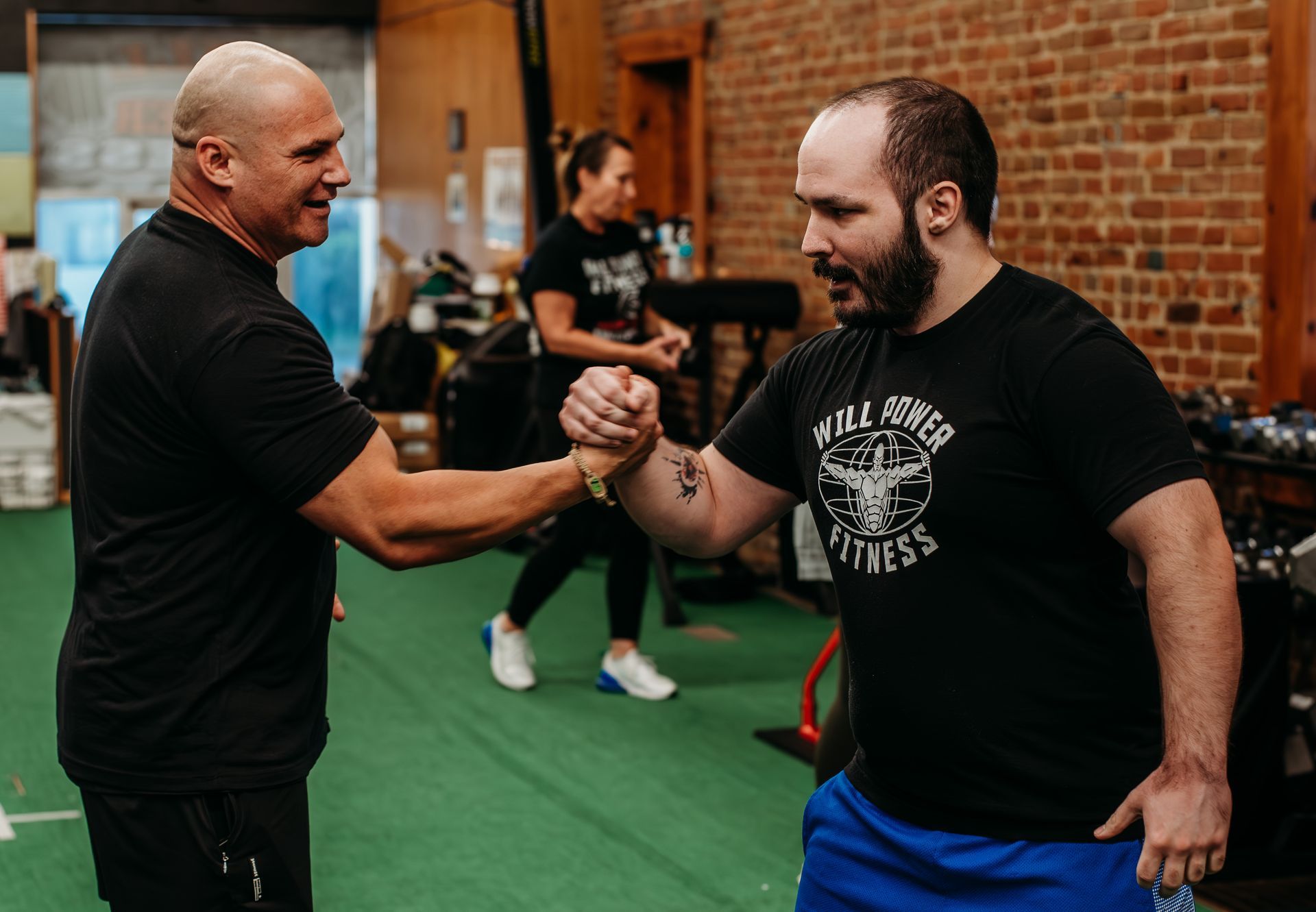 Two men fist bump in a gym. One is in a black shirt, the other in a black tee, near a brick wall.