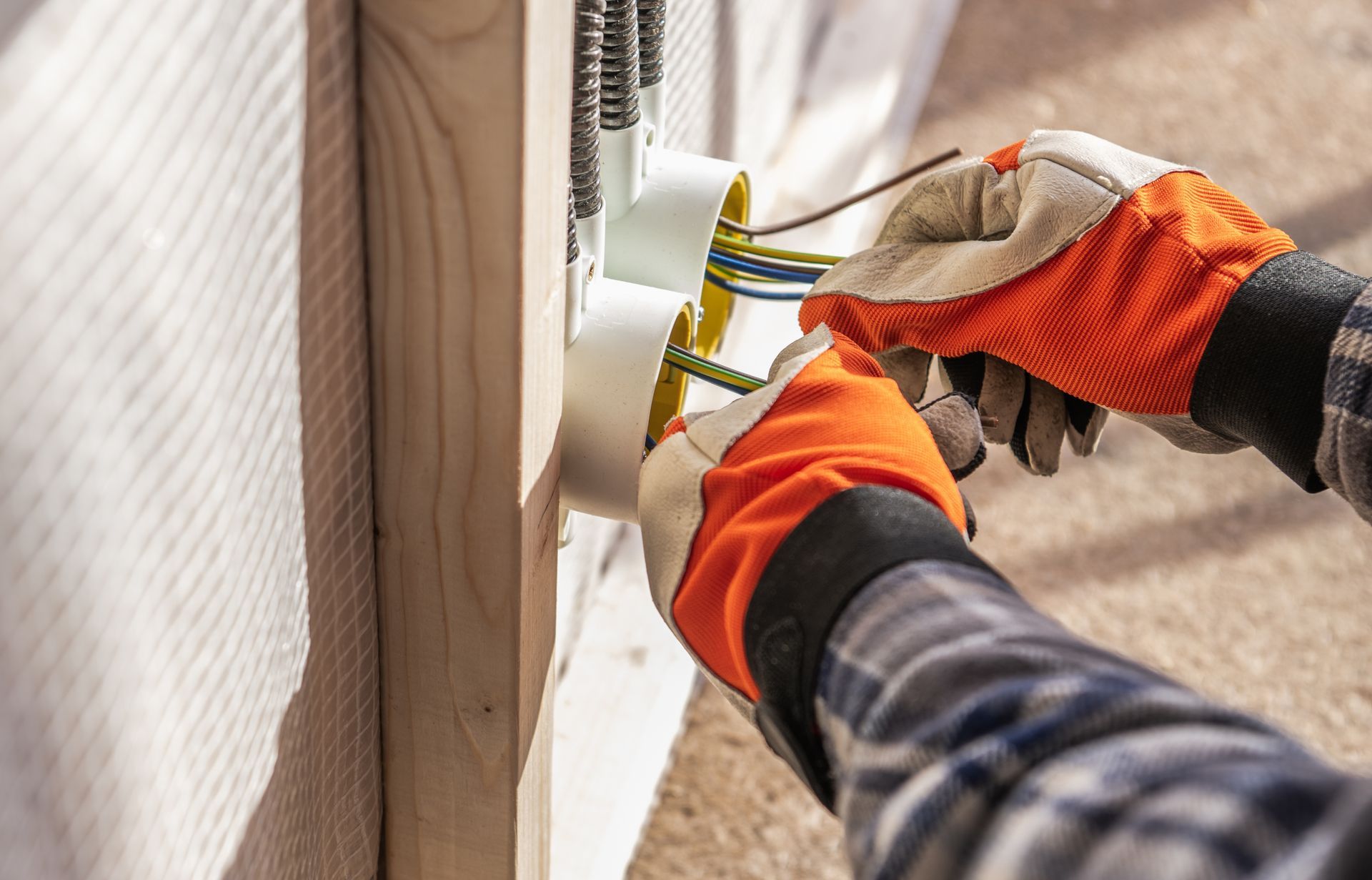 An electrician wearing orange and tan work gloves pulls electrical wires through a junction box mounted to a wood stud.