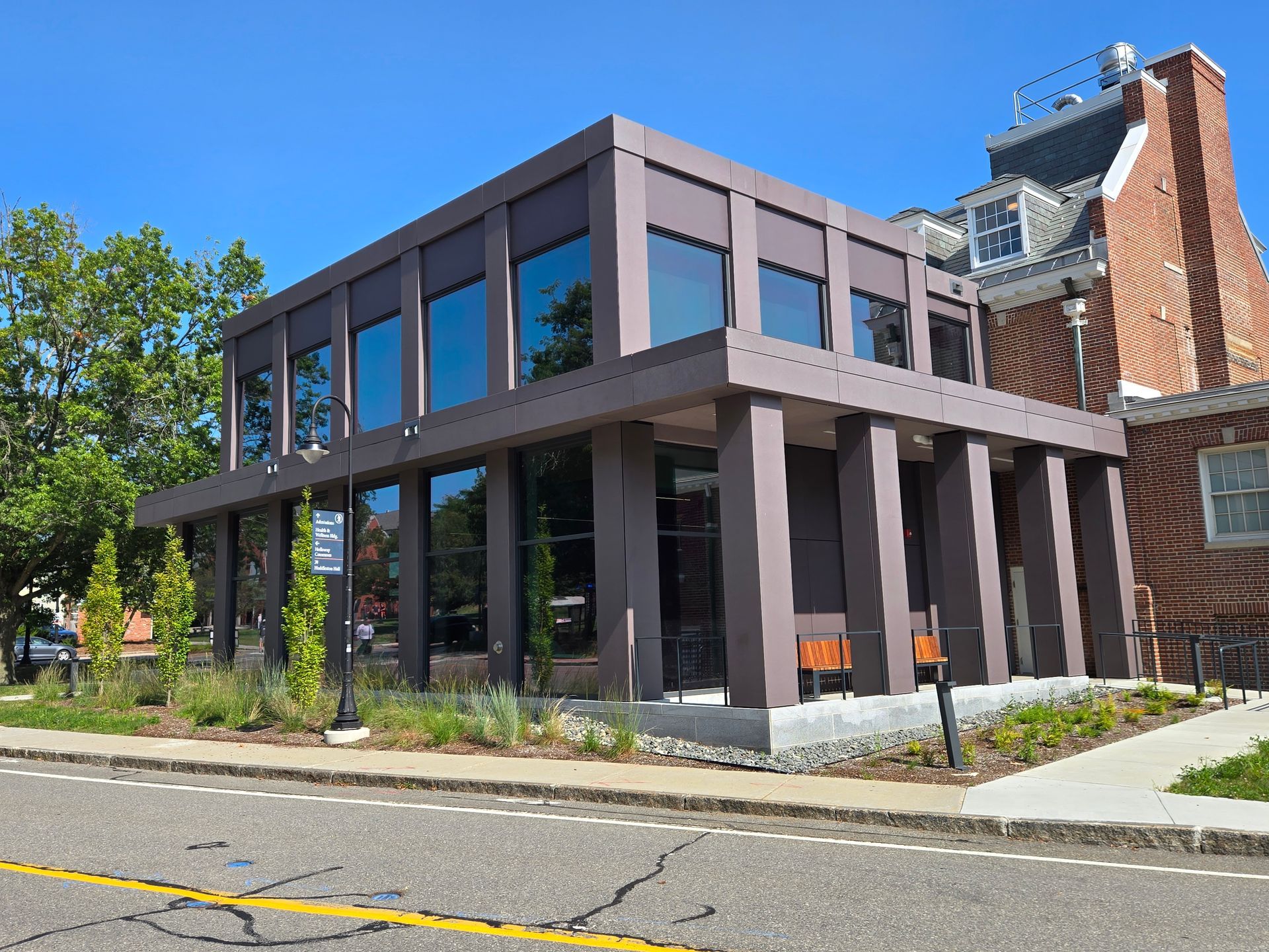 Modern brown building with large windows, next to a brick building, on a sunny street.
