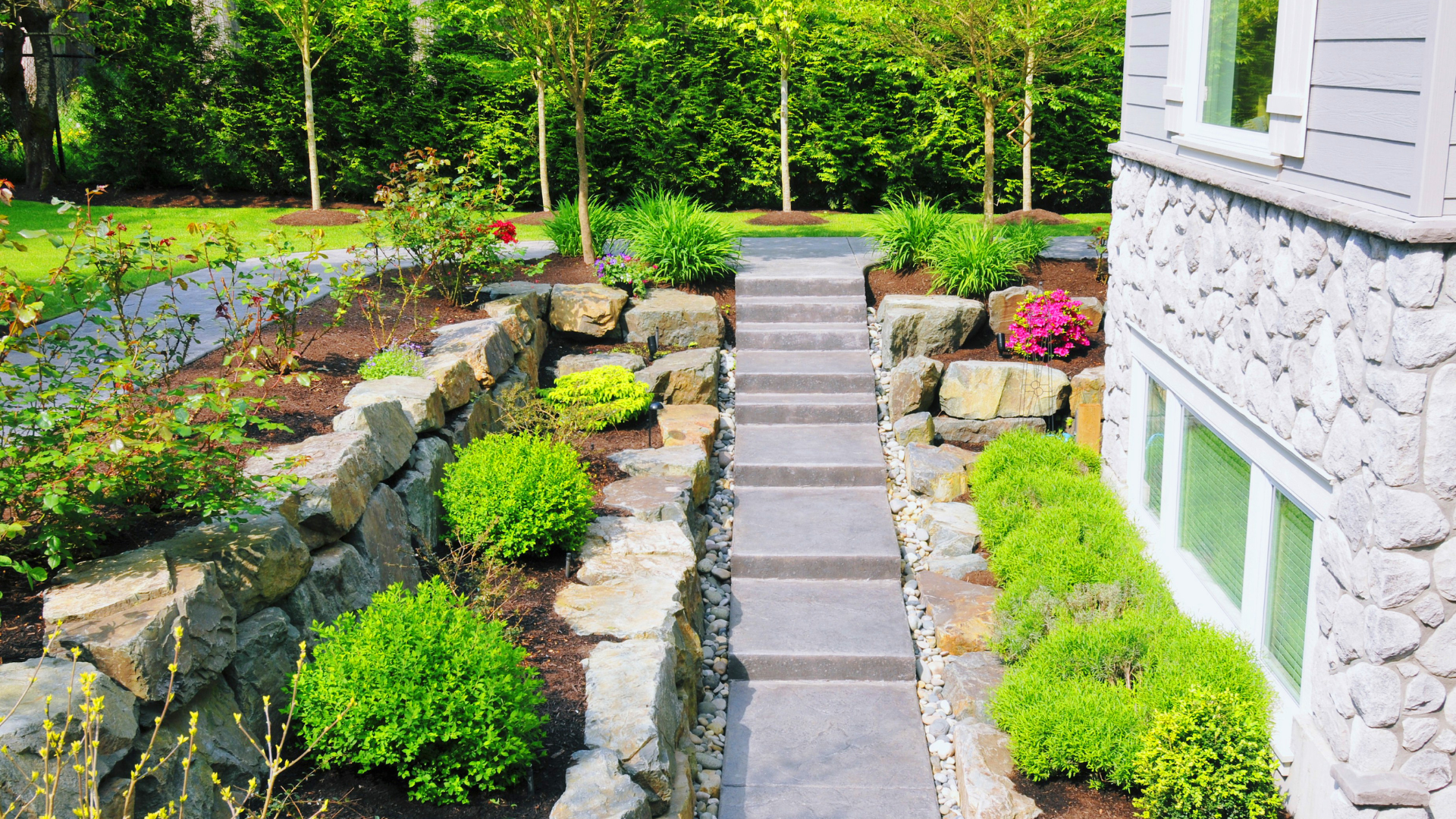 A tiered stone patio with a water feature, lounge chair, gas fire, and landscaped flower pots at dusk.