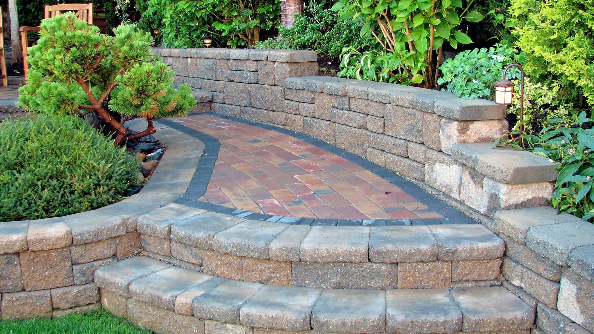 A stone patio with a rectangular swimming pool, a water feature, a waterslide tunnel, and tiered landscaping.