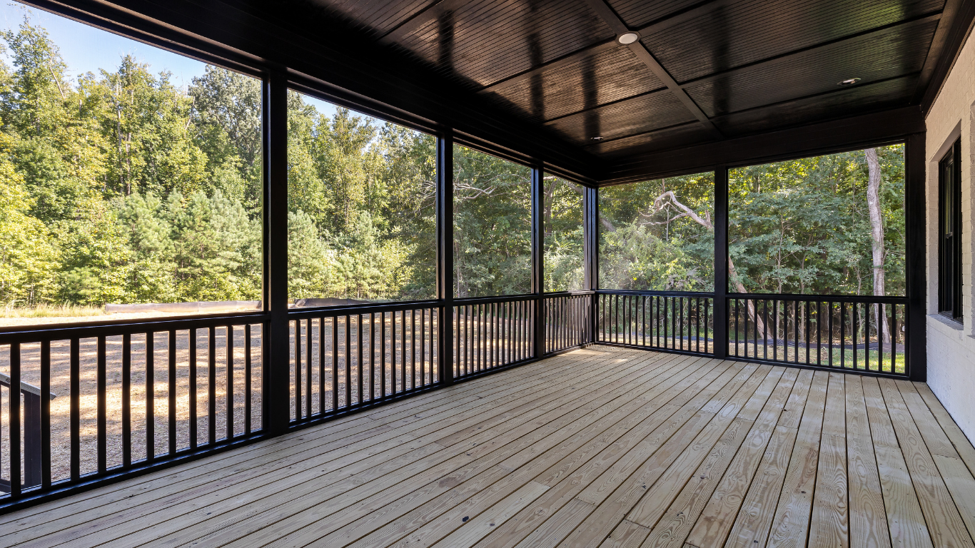 An empty screened in porch with a wooden deck and black railing.