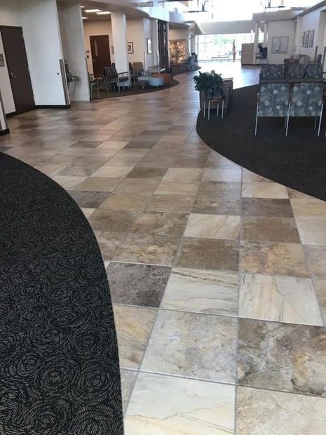 Empty hotel lobby with tan tiled floor, dark carpeted seating areas, and lounge chairs along the walls