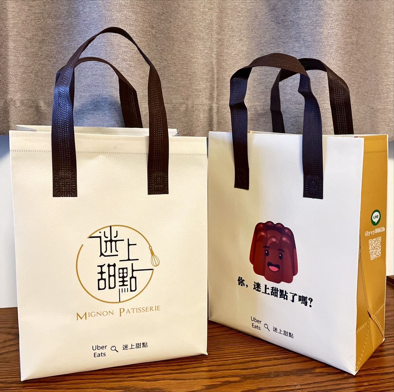 木桌上擺放著兩個米色禮品袋，袋子配有深棕色提手，其中一個印有中文，側邊為黃色。