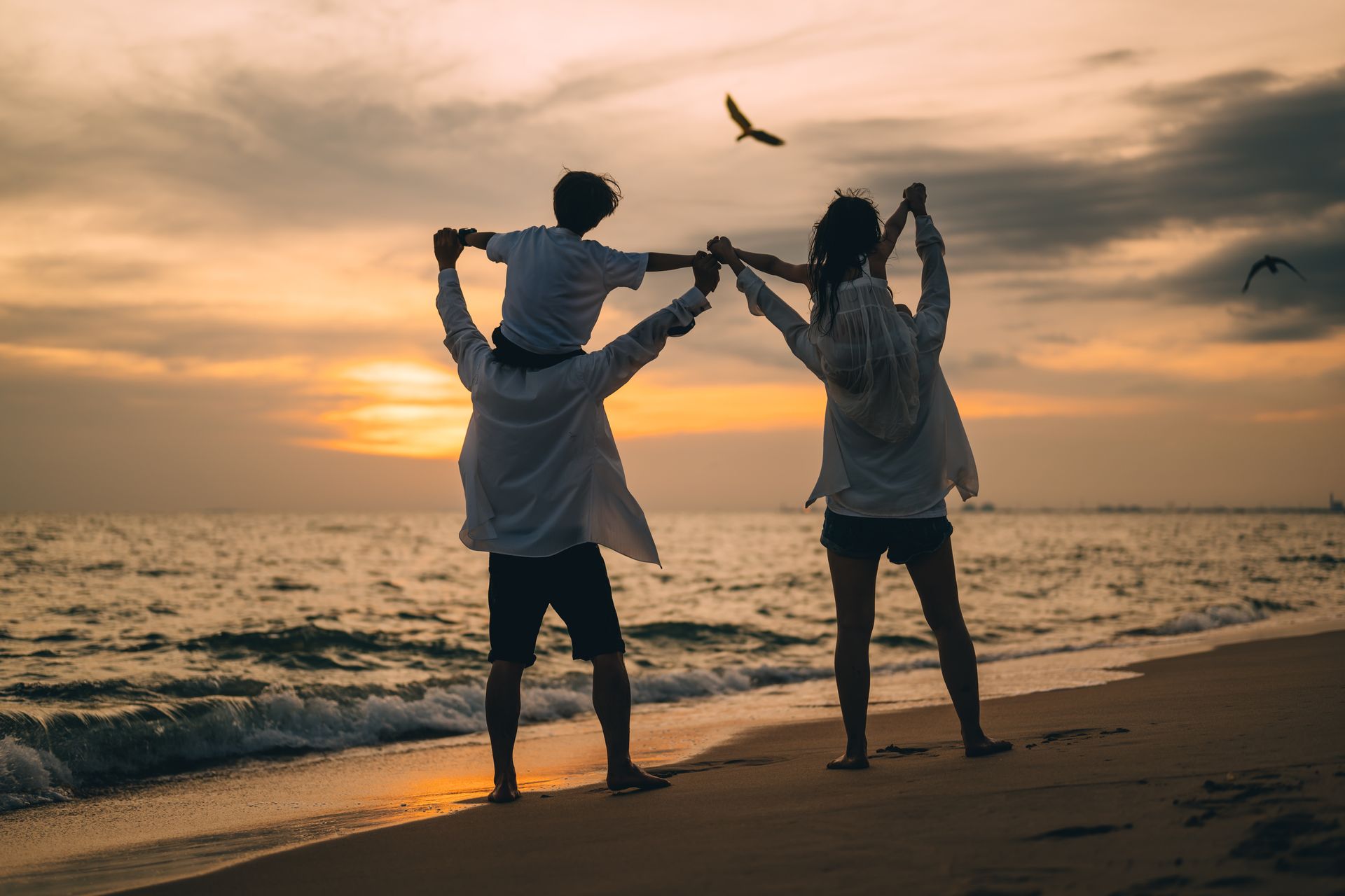 Two people on a beach at sunset, holding hands and jumping, with waves and birds in the sky.