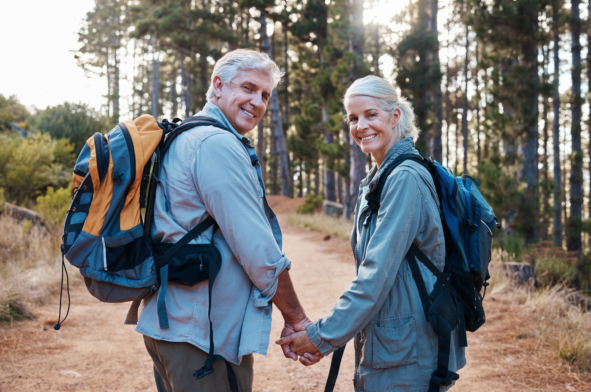 Two hikers with backpacks smiling and holding hands on a forest trail at sunset