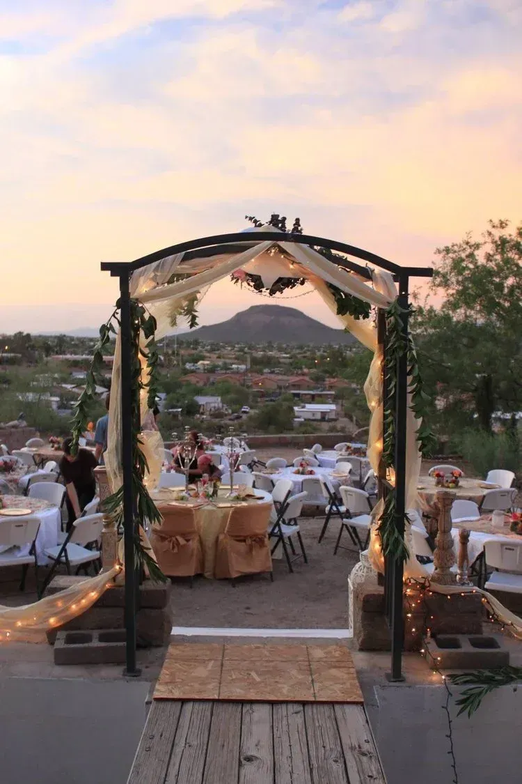 Wedding reception setup with archway overlooking a desert landscape at sunset. Tables set, some with gold linens.