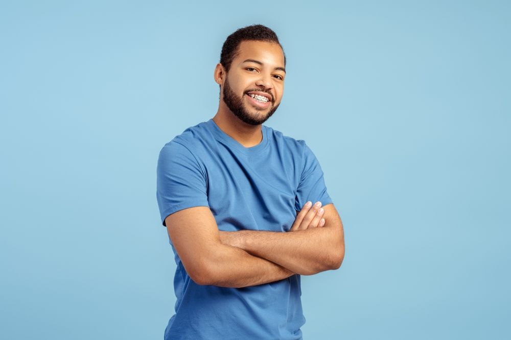 A man with dark skin and a beard in a blue shirt smiles with arms crossed against a blue backdrop.