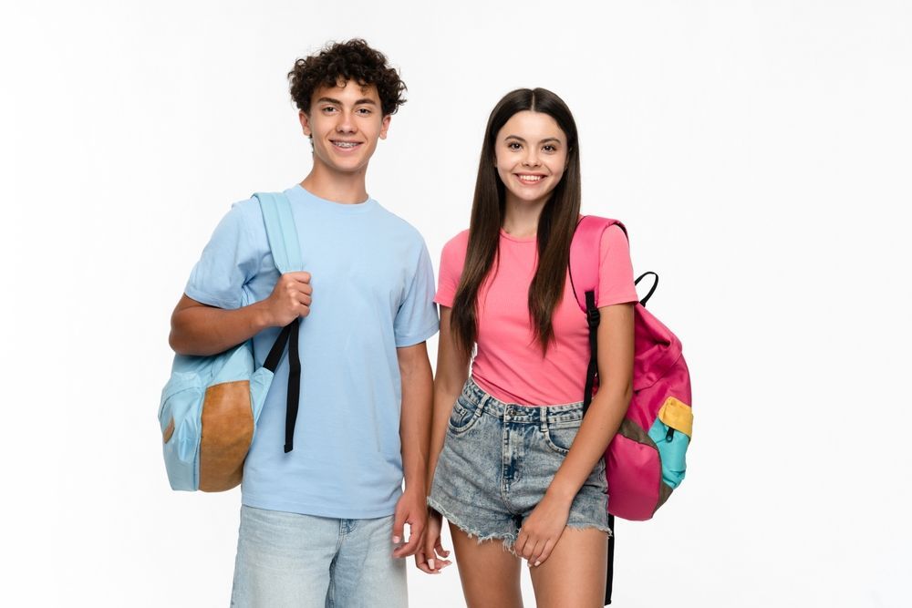 Two smiling teens with backpacks, boy in blue shirt and girl in pink, against a white background.