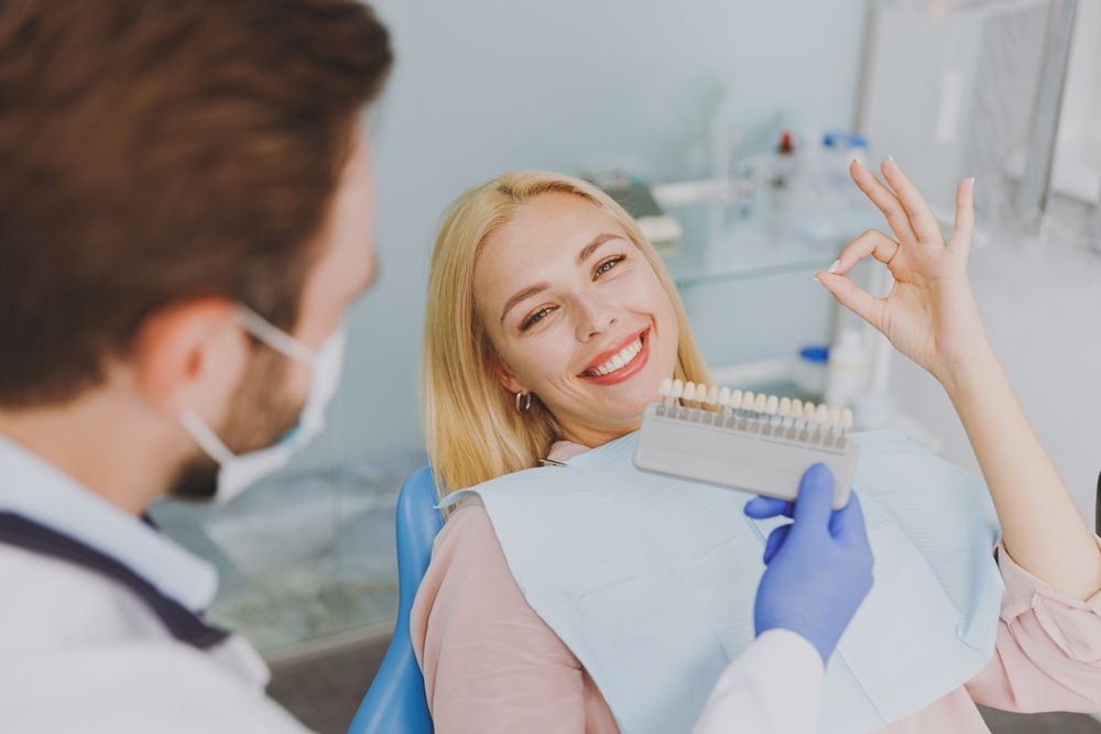 A woman is sitting in a dental chair while a dentist shows her a tooth color chart.