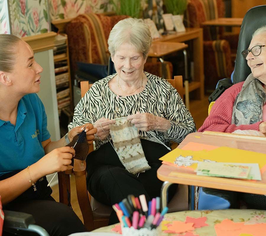 Caregiver assists elderly woman knitting, another woman beside her smiles. Indoors, crafting materials present.