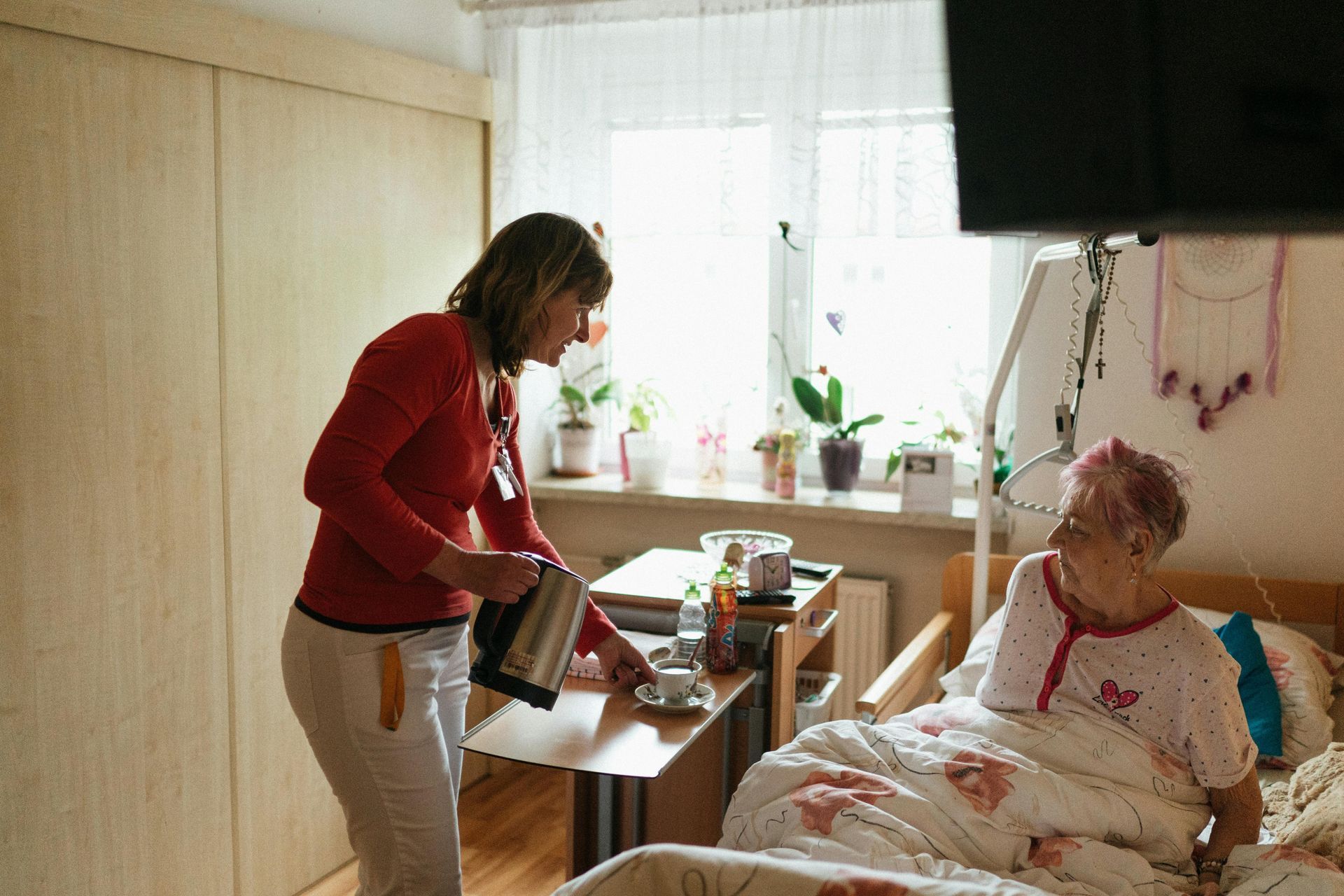 Caregiver pours water from a kettle into a cup for a person in bed in a room with a window.
