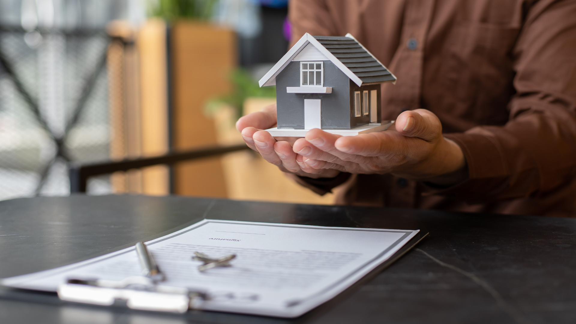 A person holds a small model house above a desk with documents and a pen, representing a real estate or mortgage process.