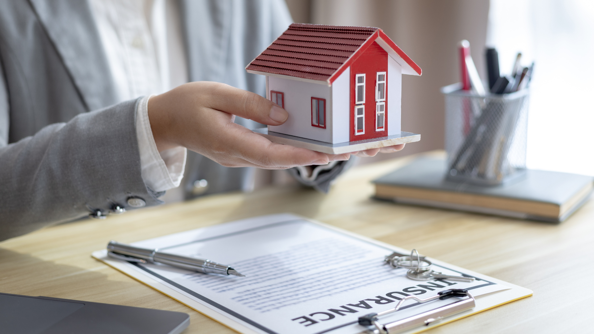 A person in a business suit holds a model house over a desk with insurance paperwork and a pen.