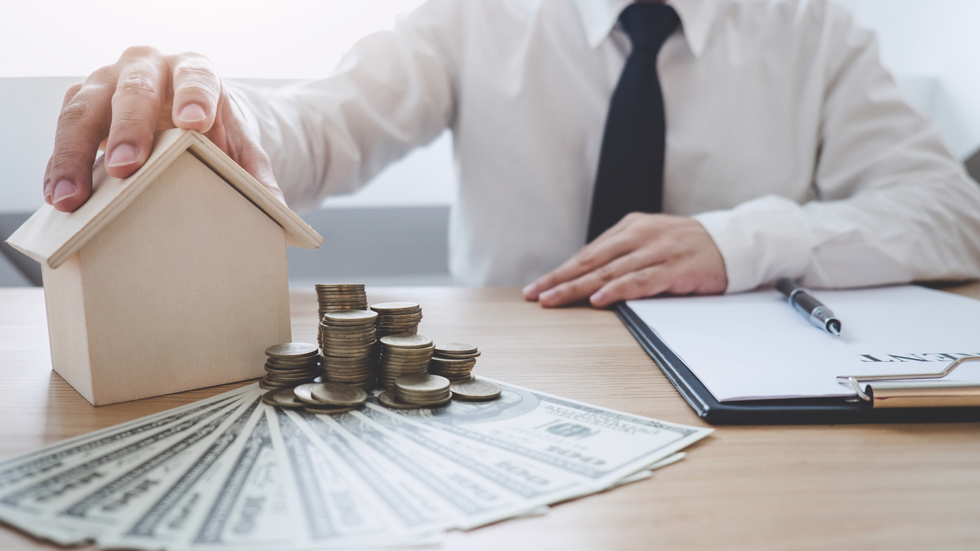 A person in a shirt and tie sits at a desk with a model house, a stack of coins, and a fan of dollar bills.