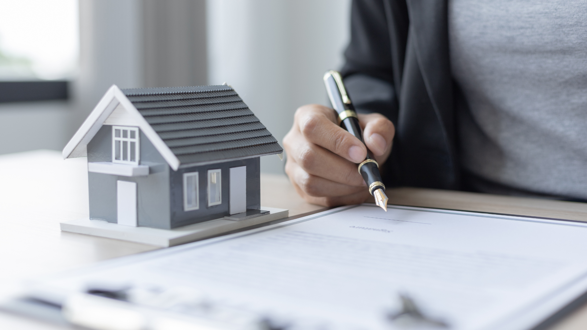 A person in a business suit signs a document next to a small model house on a desk.