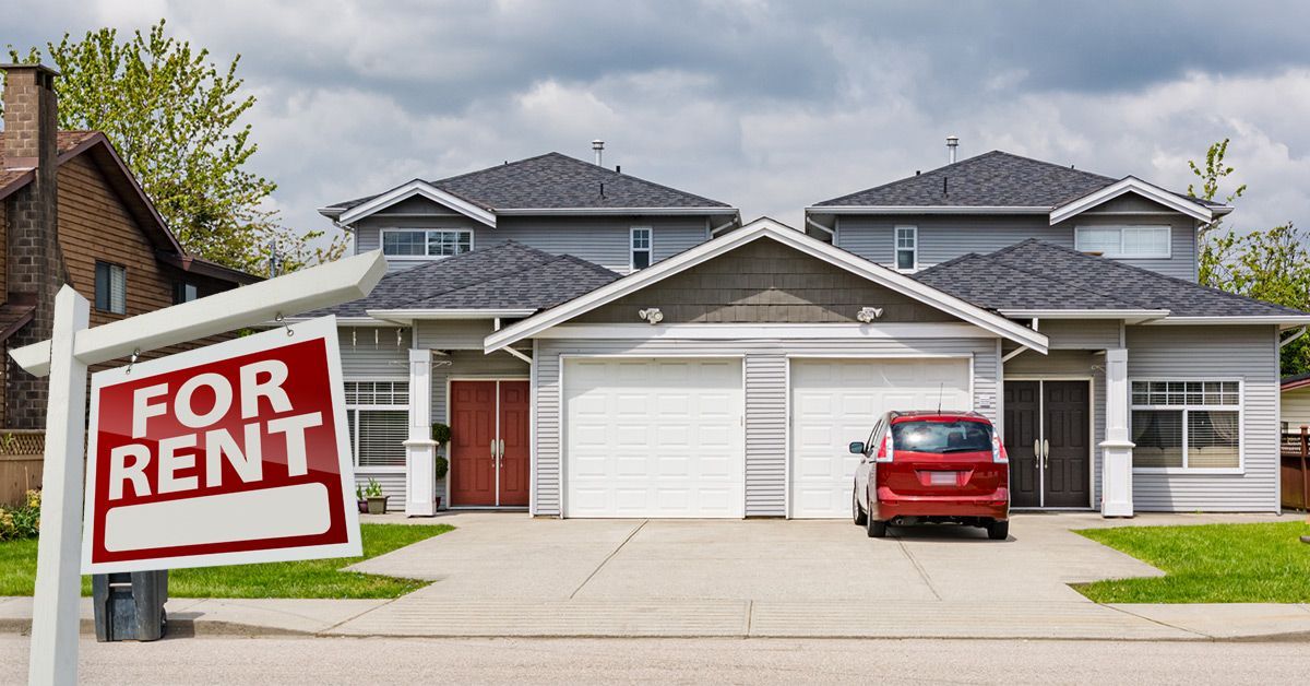 A person in a light shirt holds a calculator and pen next to a model house on a desk with a clipboard.
