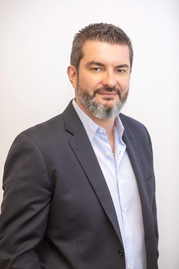 A man with a beard, wearing a pinstriped navy blazer and a light blue dress shirt, stands against a white background.