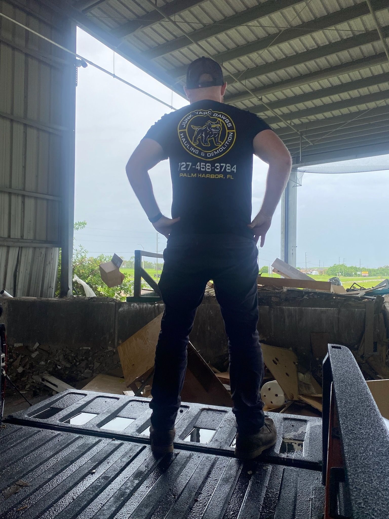 Man in black shirt and pants standing in a truck bed, hands on hips, near debris. Inside a shed.