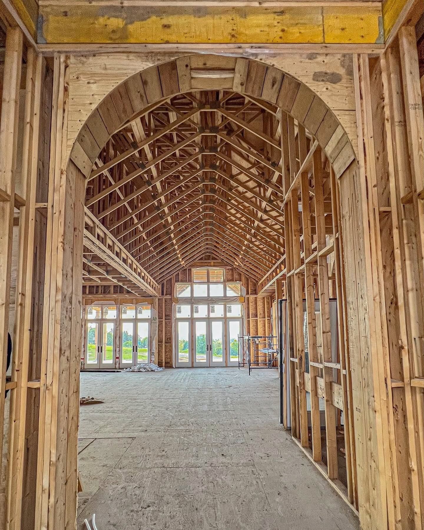 Interior view of a building under construction, framed with wood. Arched doorway leads to a long room with windowed walls.