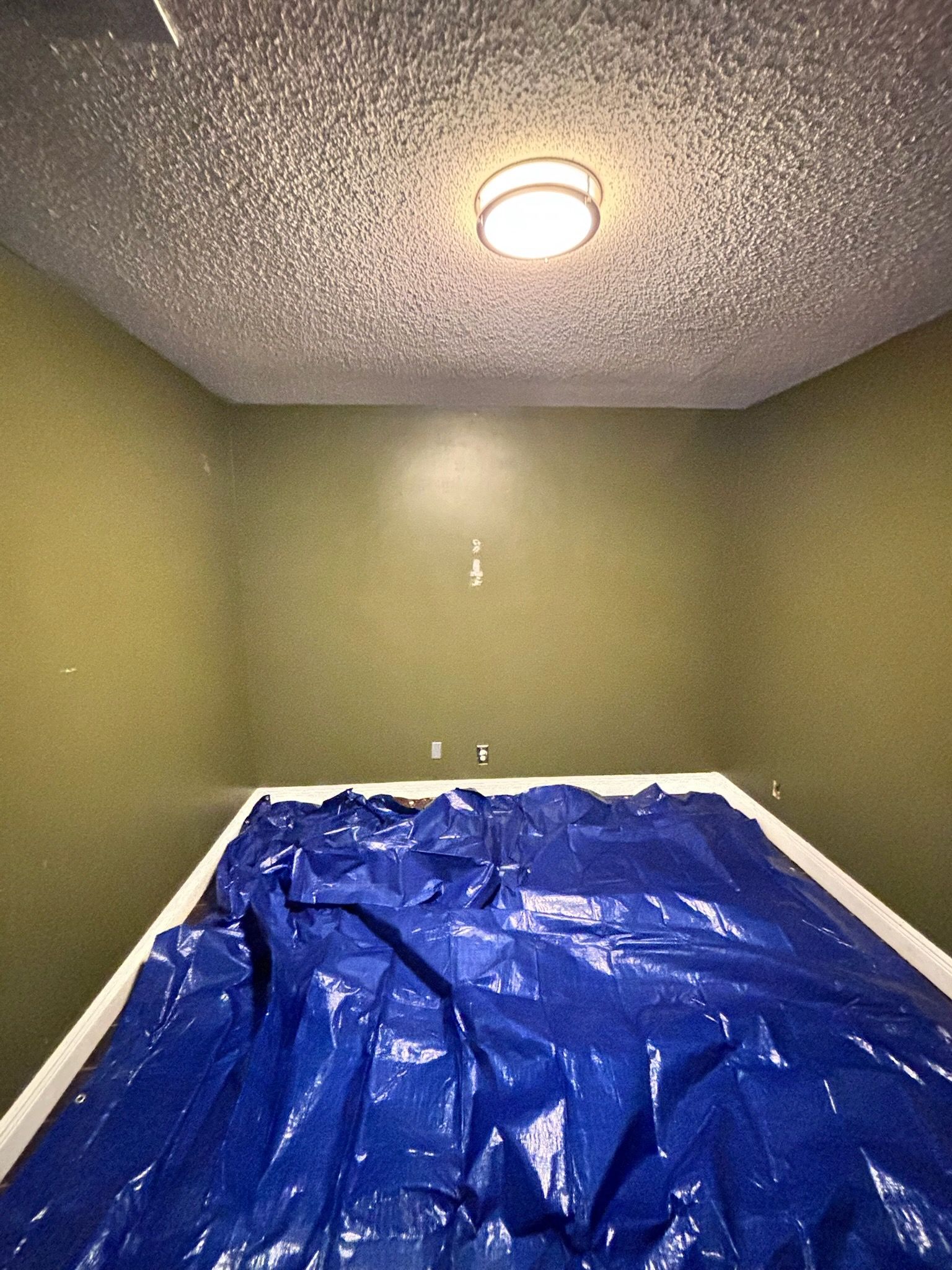 Empty room with blue tarp covering the floor, olive green walls, popcorn ceiling, and a central light fixture.