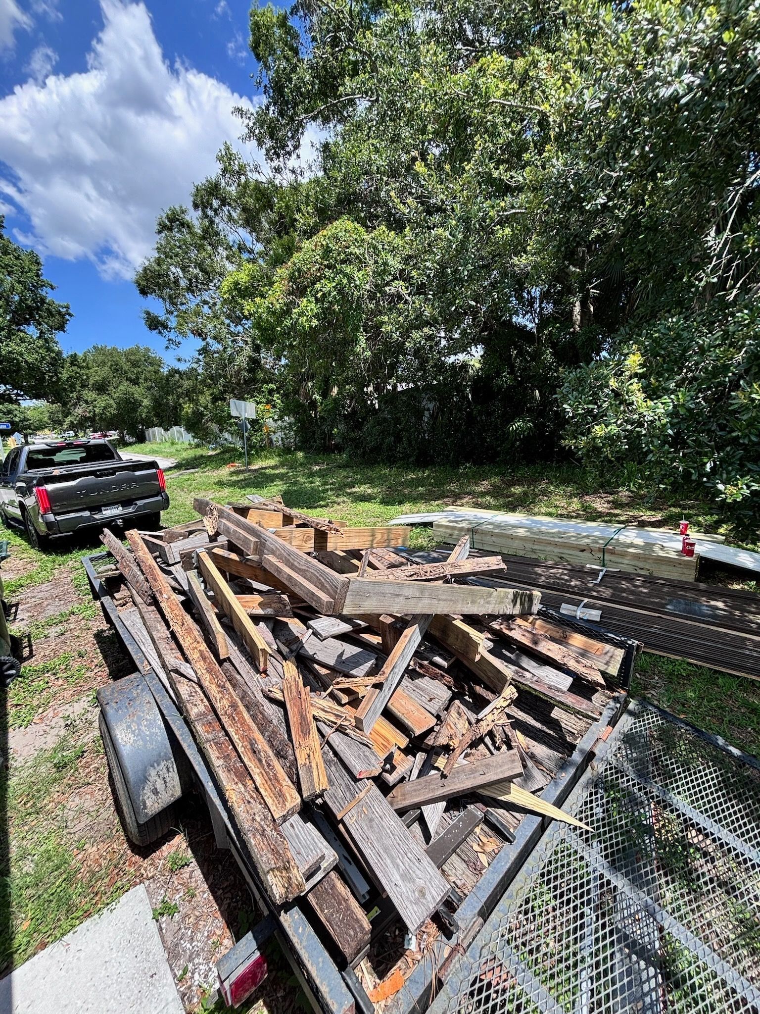 Trailer loaded with weathered wooden planks, parked next to a truck in a grassy area with trees and blue sky.