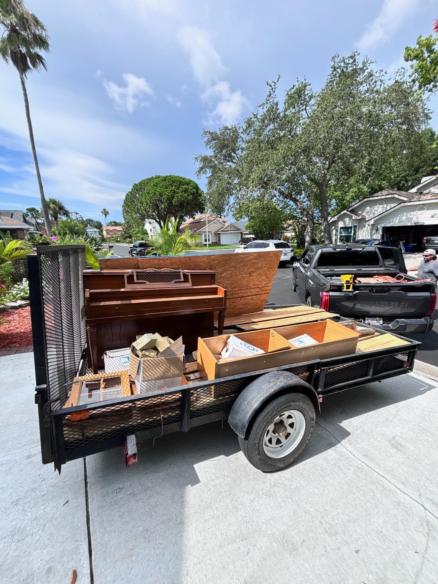 A trailer loaded with discarded furniture and items is parked in a driveway on a sunny day.
