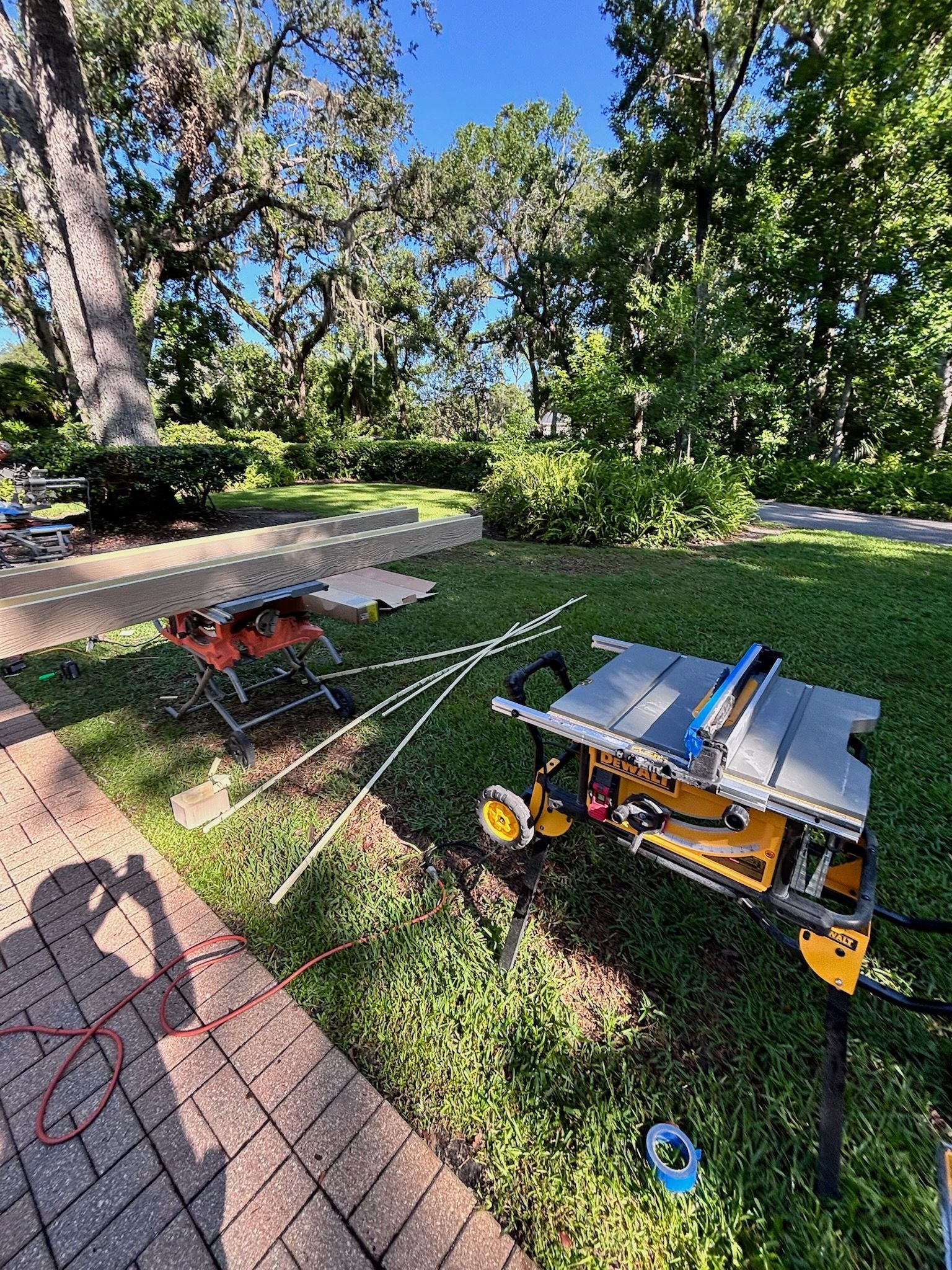 Two portable table saws on grass outdoors; one orange, one yellow/gray. White cords connect them.