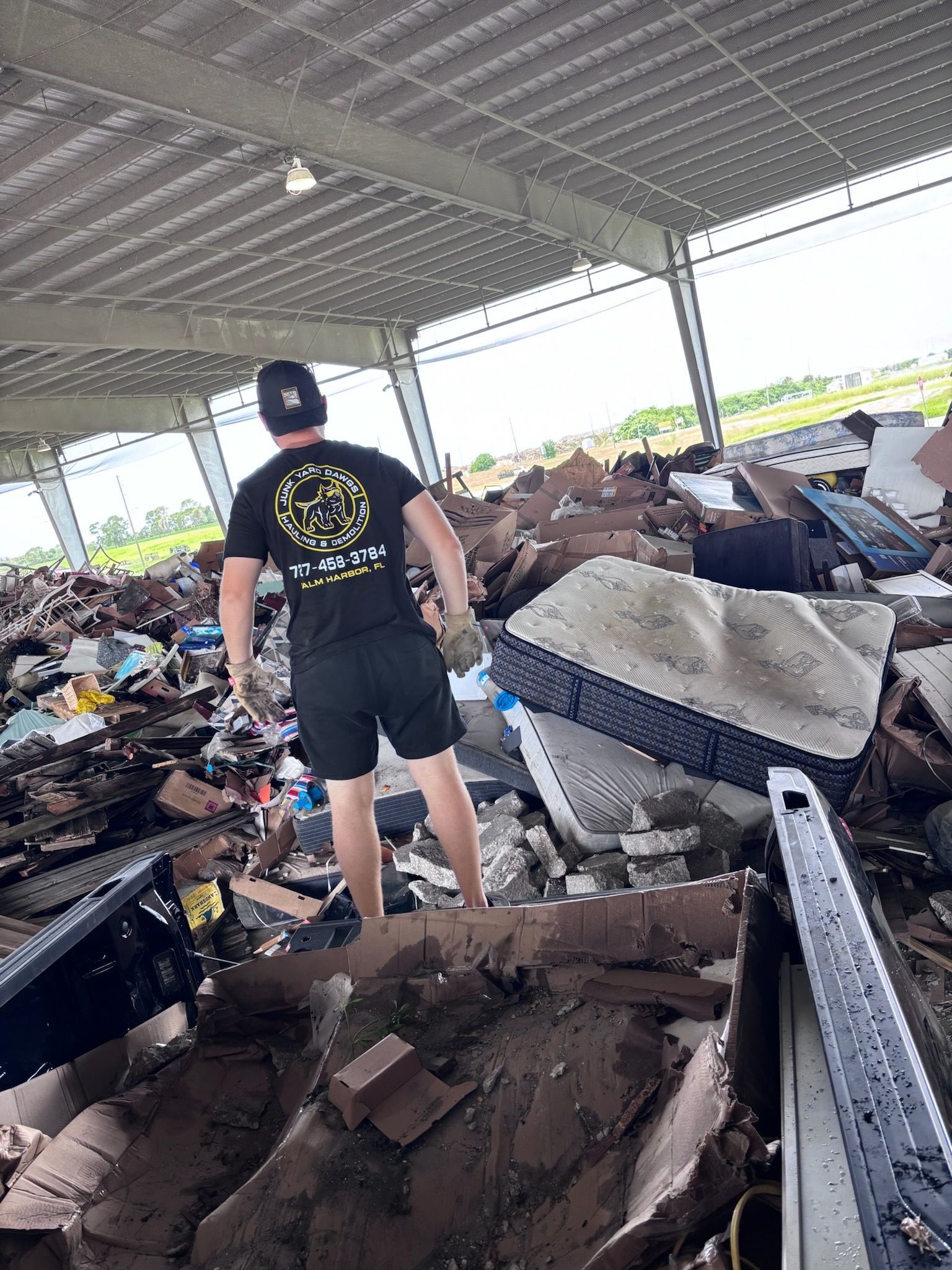 Person in black shirt and shorts standing on top of a pile of trash inside a large, open-air building.