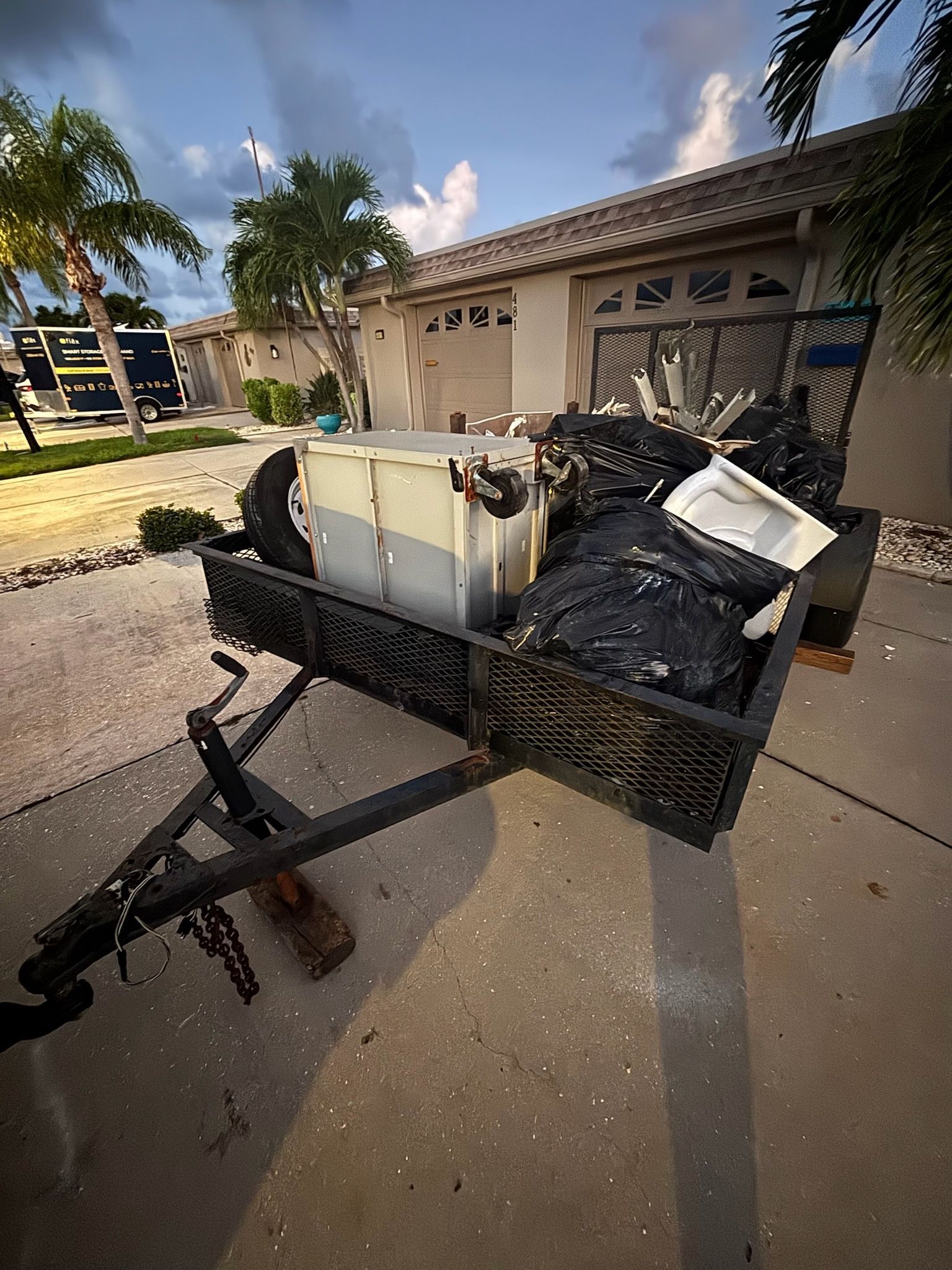 A trailer loaded with trash bags, a white box, and other debris, parked on a concrete driveway near a house and palm trees.