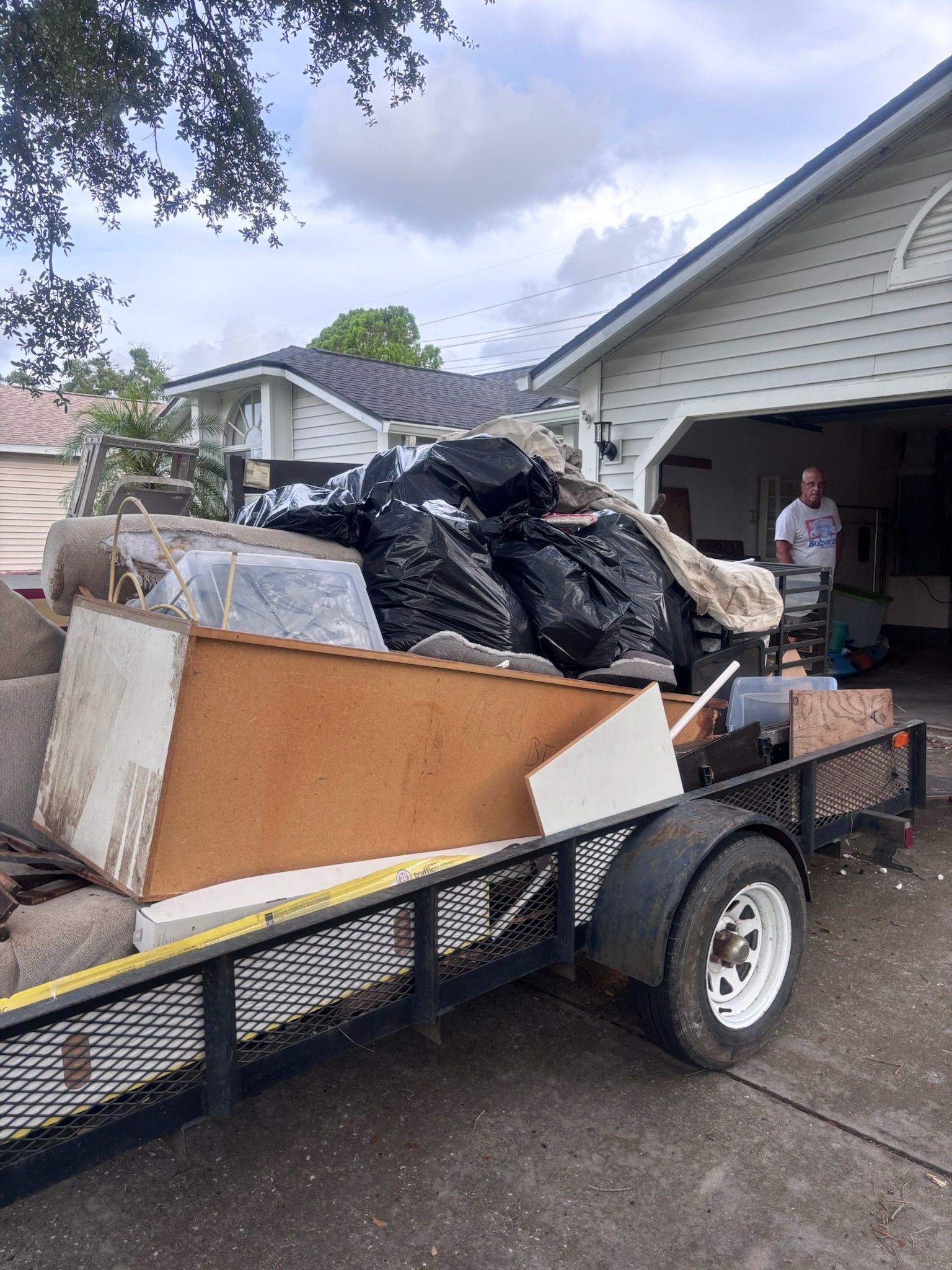Trailer loaded with household debris parked in front of a house. A person stands nearby, looking on. Overcast day.