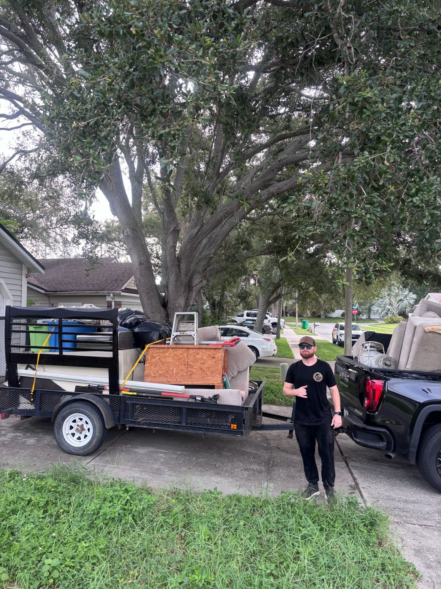 Person stands next to a truck with a trailer loaded with furniture. Outdoors in front of a house.