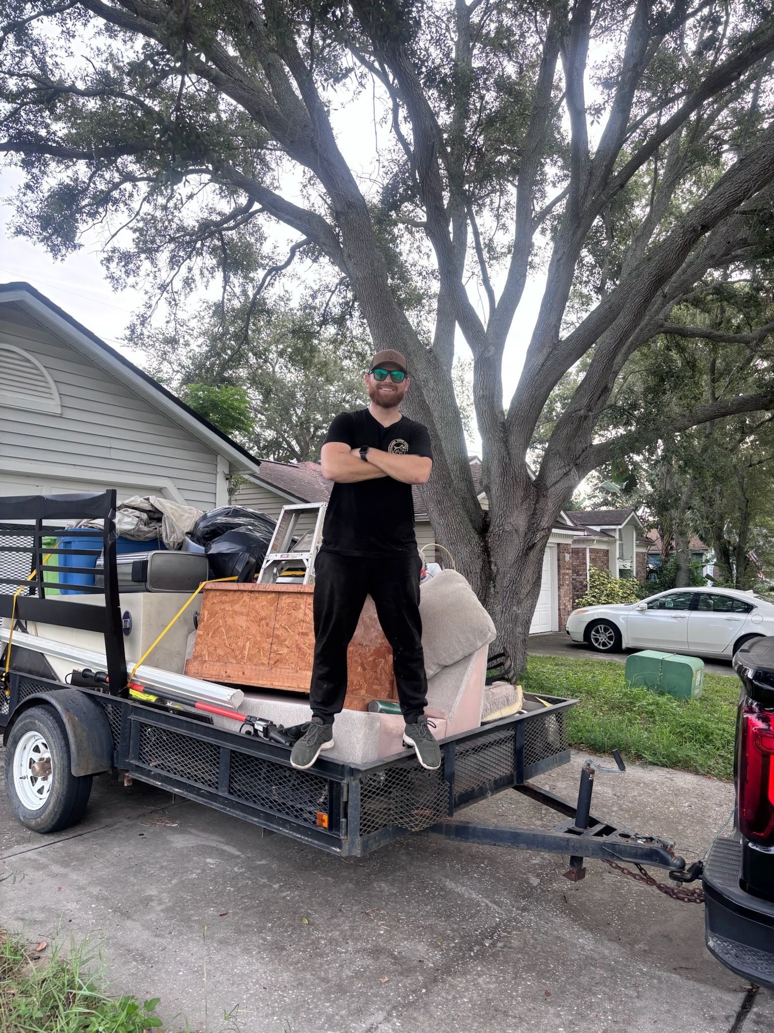 Man standing on loaded trailer, arms crossed. Outdoors, residential setting.