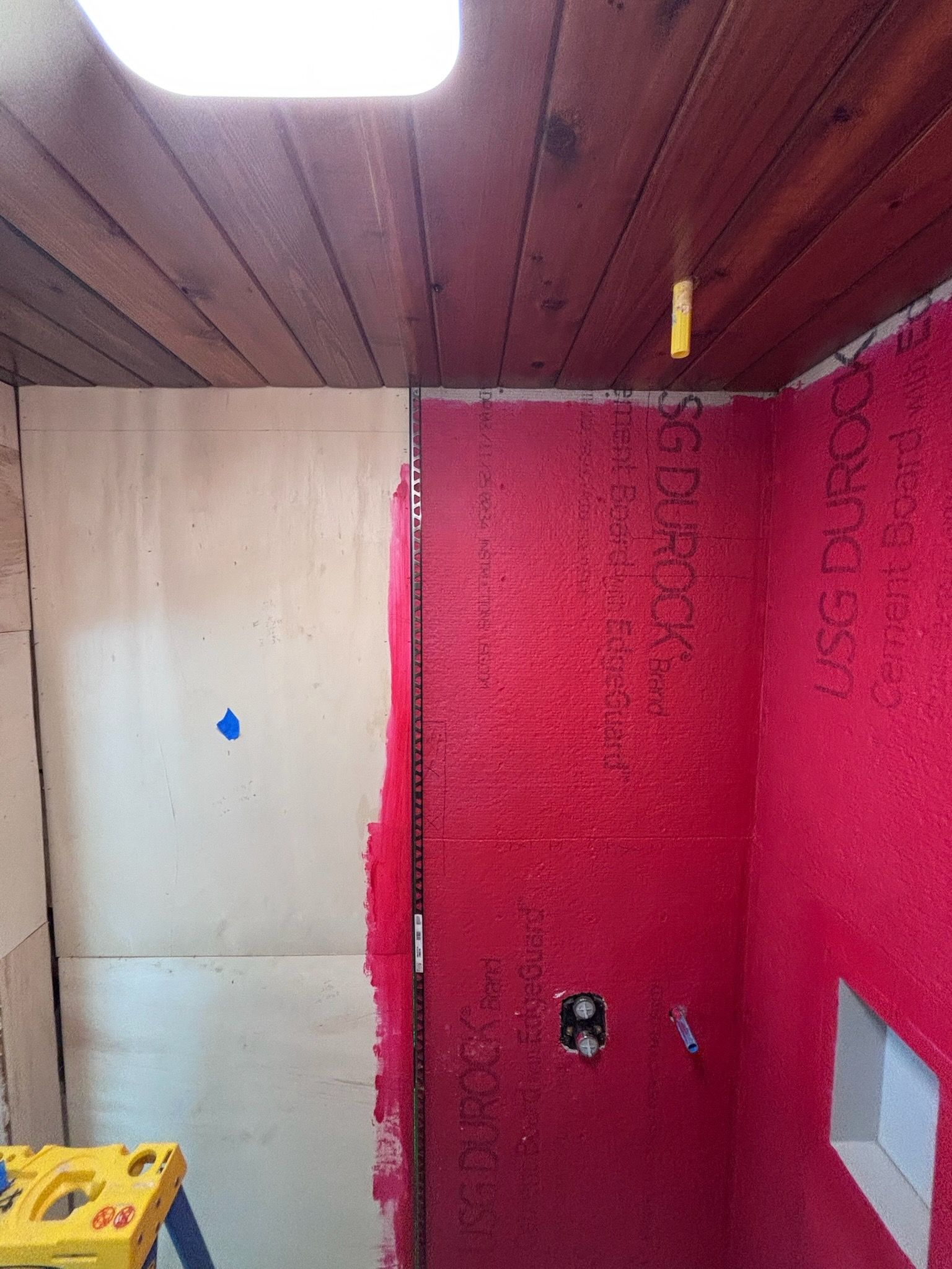 Interior view of a shower under construction, red waterproof membrane on the wall, wood ceiling.