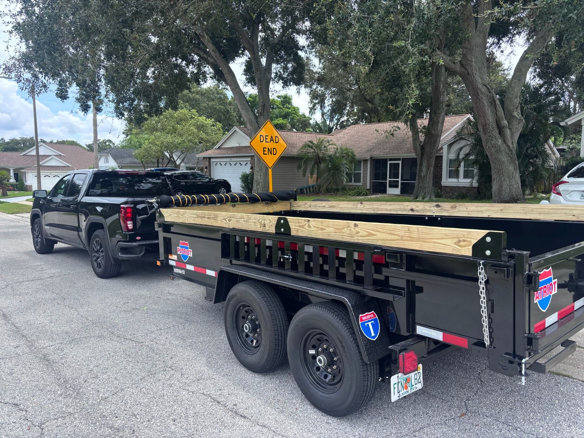 Black truck towing a black trailer with wood planks in a residential street, under a 