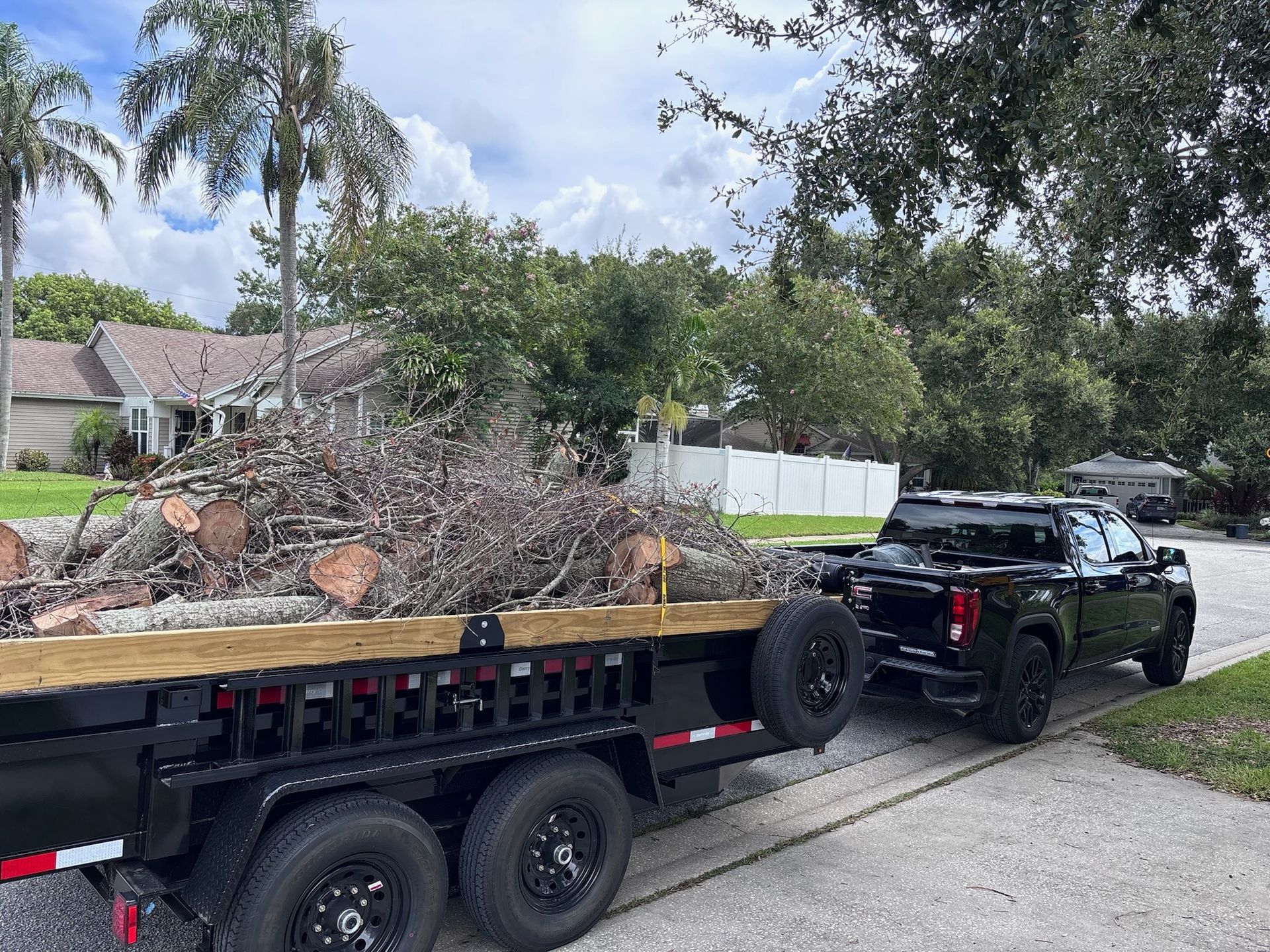Black truck towing a trailer loaded with tree branches on a residential street.