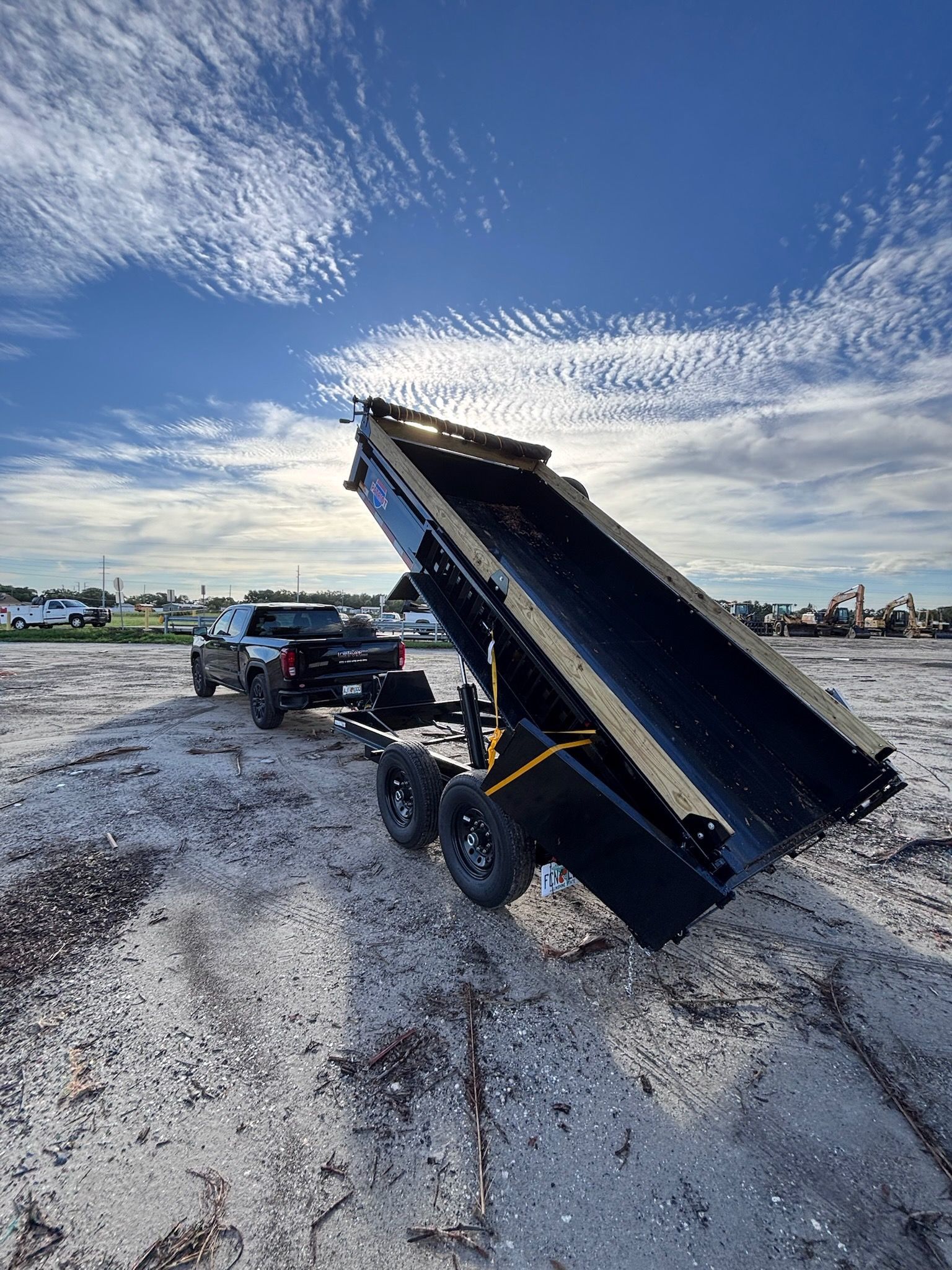 Black pickup truck towing a black dump trailer, tilted up in an open field under a blue sky.