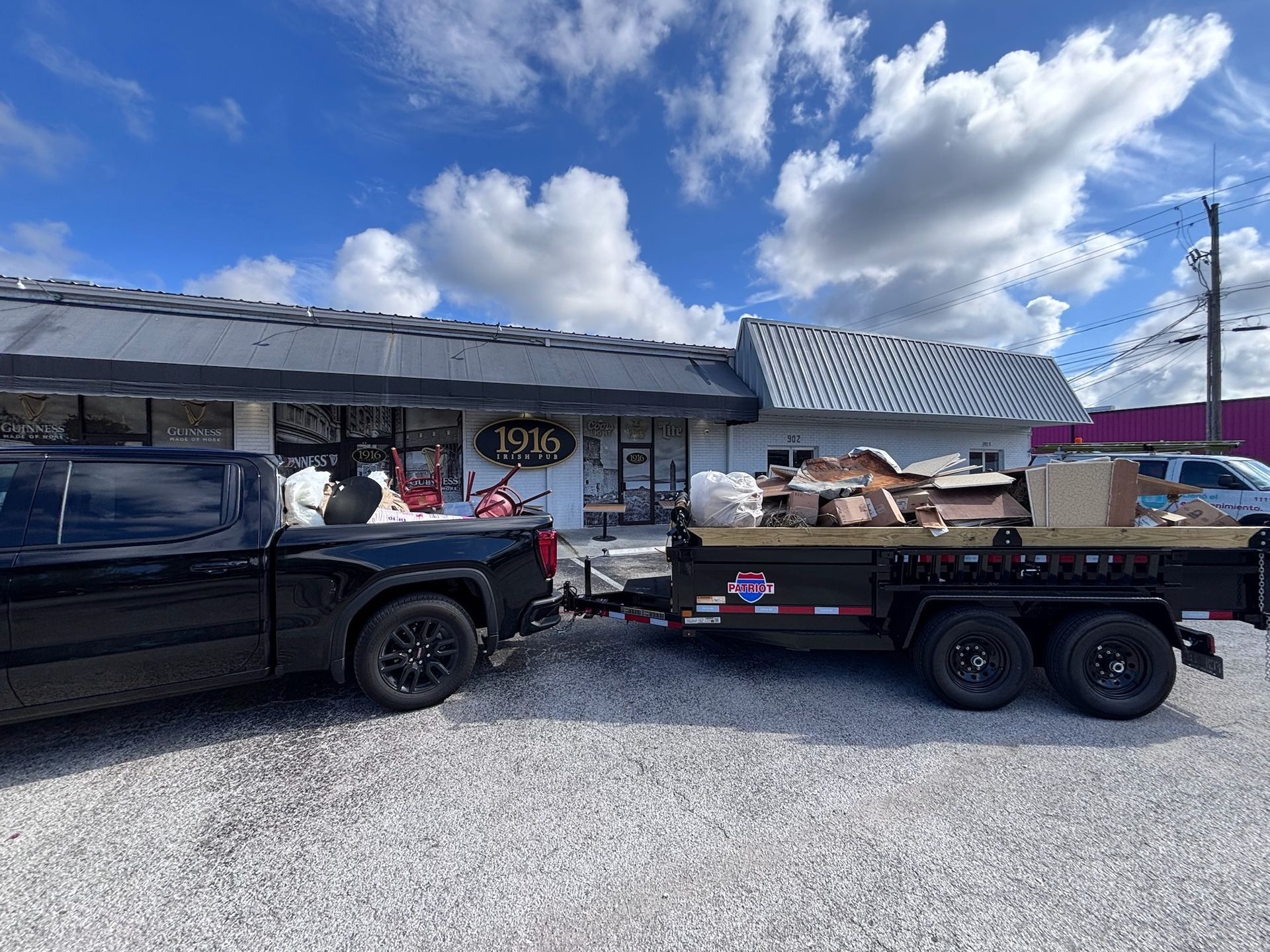 Black truck towing a trailer loaded with debris in front of a store with a sign.