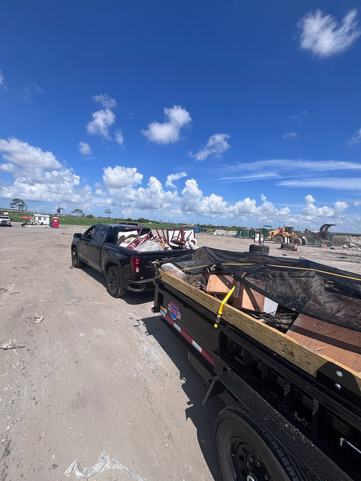 Black truck towing a trailer loaded with debris on a dirt lot under a blue sky.