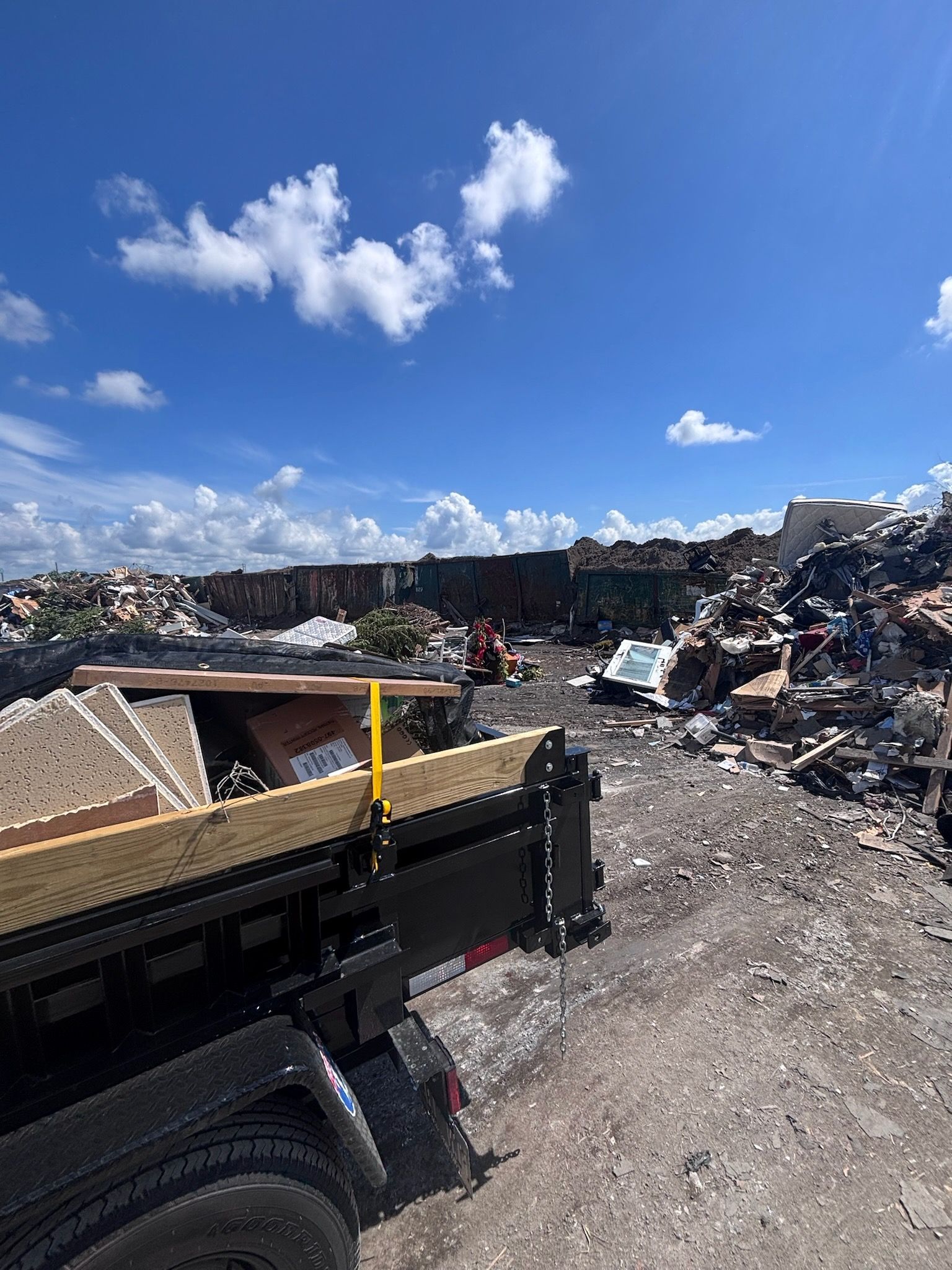 A black trailer filled with debris in a junkyard under a bright blue sky with scattered clouds.