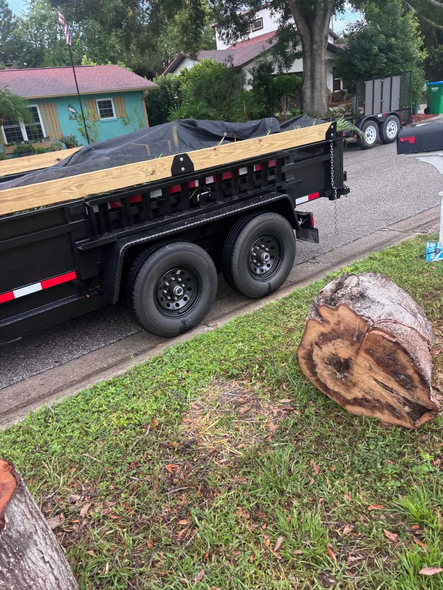 Black dump trailer parked on a residential street next to a tree stump and grass.