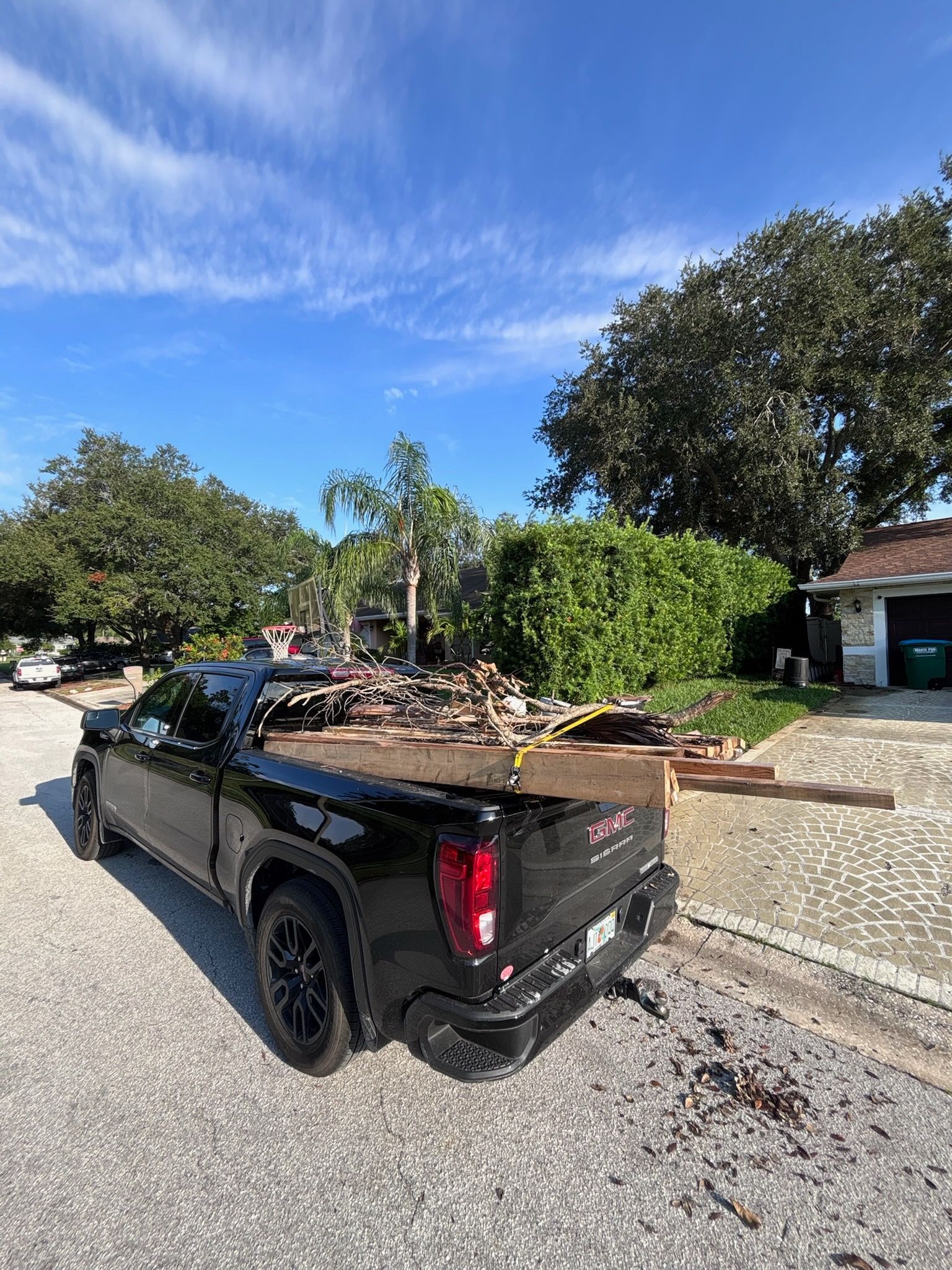 Black pickup truck loaded with lumber parked on a street; trees and house in background.