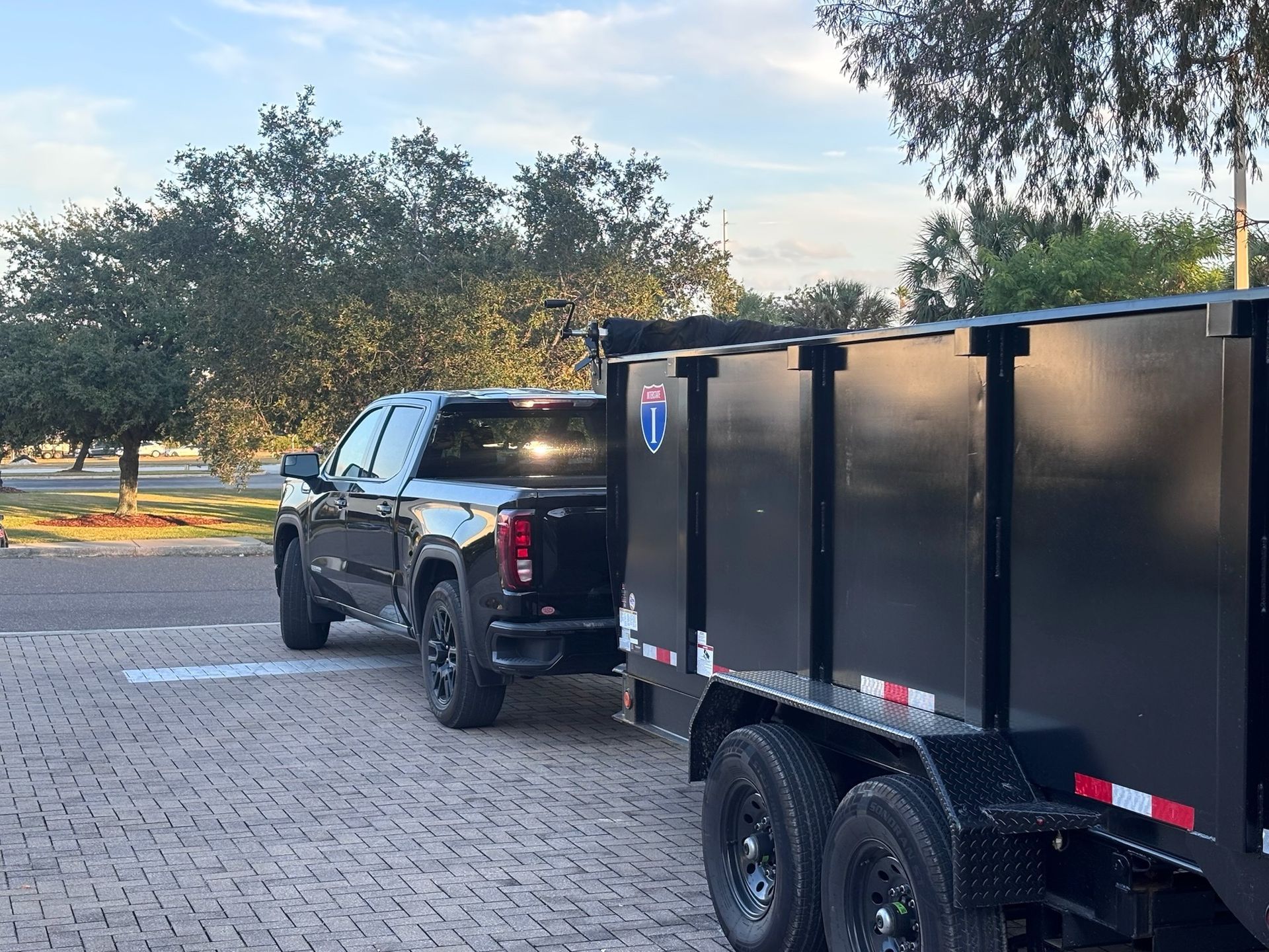 Black pickup truck towing a black dumpster trailer parked on a brick-paved area.
