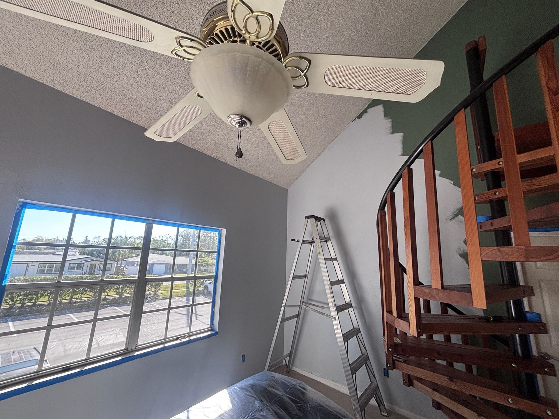 A room with a ceiling fan, a window, and a spiral staircase being painted; ladder present.
