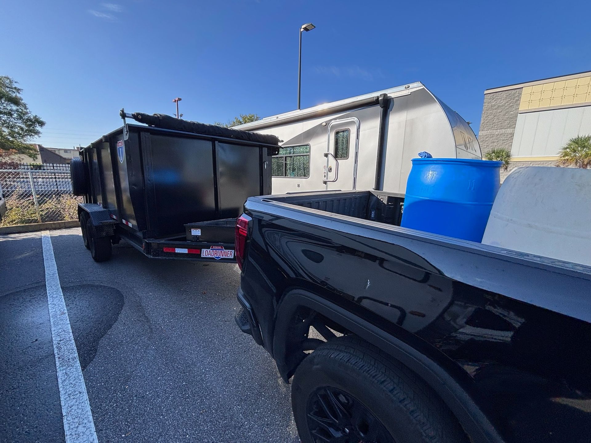 Black truck towing a black dumpster trailer in a parking lot on a sunny day. A blue barrel rests in the truck bed.