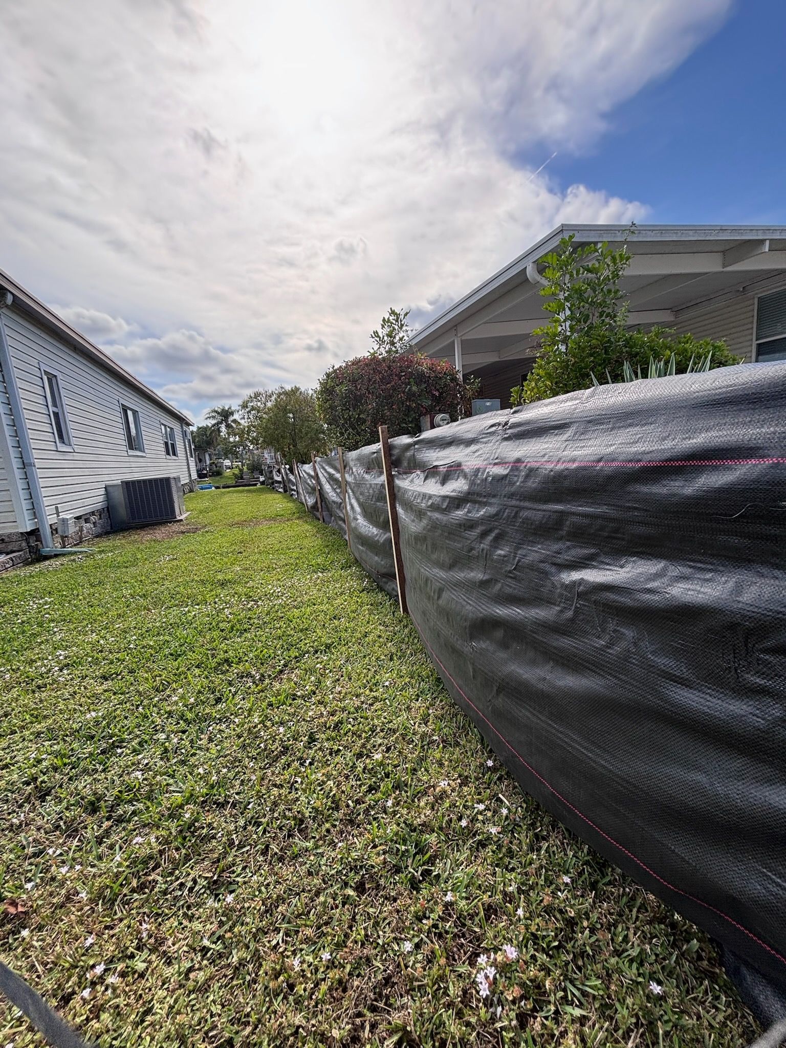 Black construction fence along a grassy yard, with houses and a partly cloudy sky in the background.
