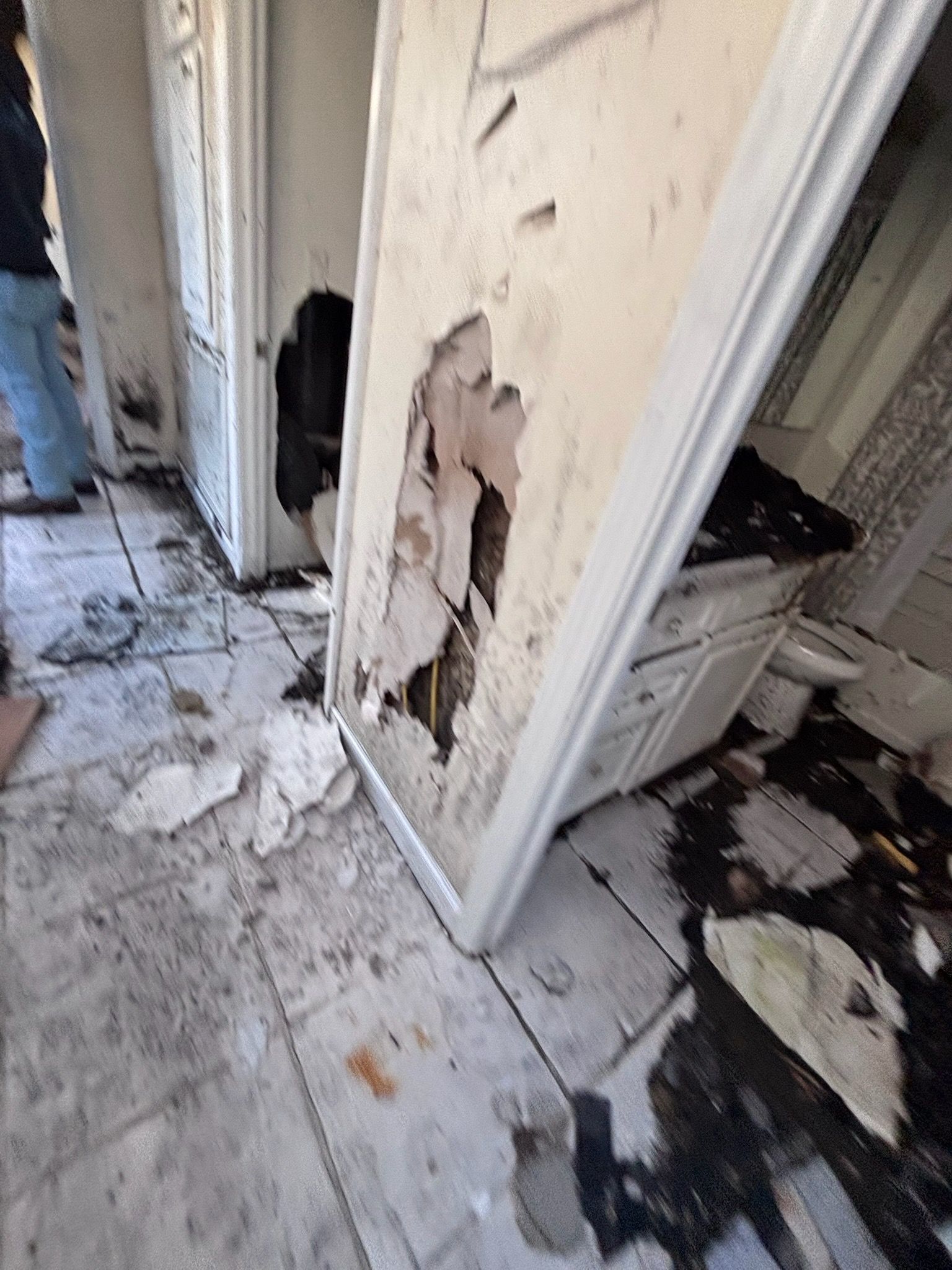 Interior damage: peeling wall, broken door, damaged floor, possible fire damage, standing person.