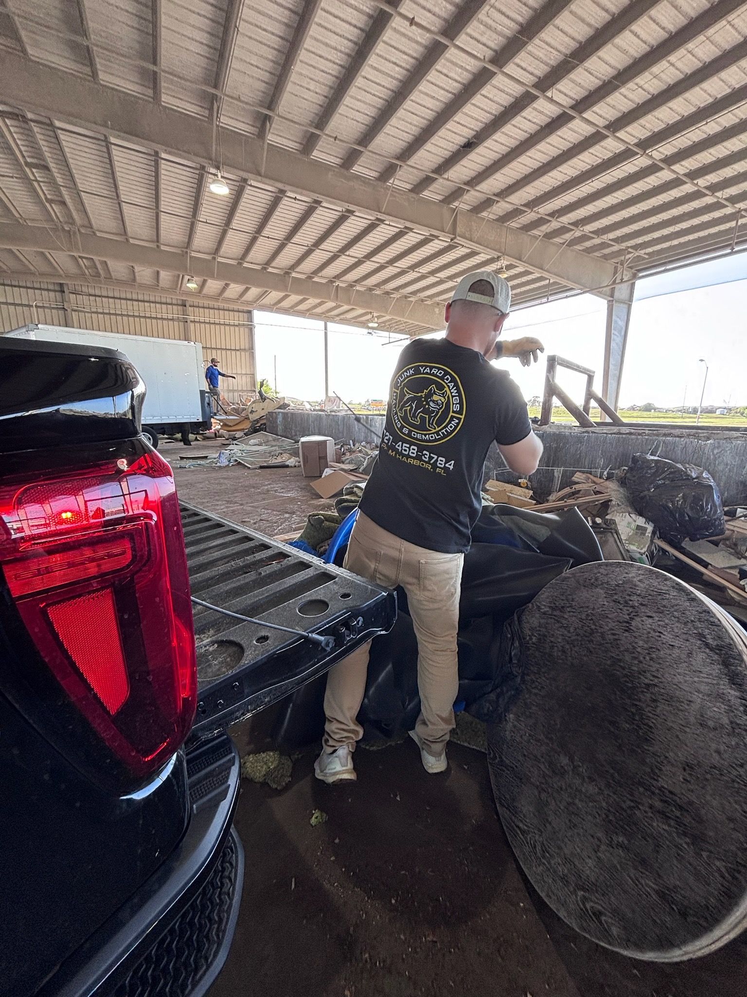 Man loading debris into truck bed in a large, open-air structure. Black truck, trash bags, tires visible.