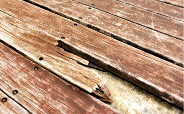 A photo of a deck that has taken a lot of damage over time from weather and natural elements. The wood is cracking and falling apart. The protective paint coating has faded from the sun. 